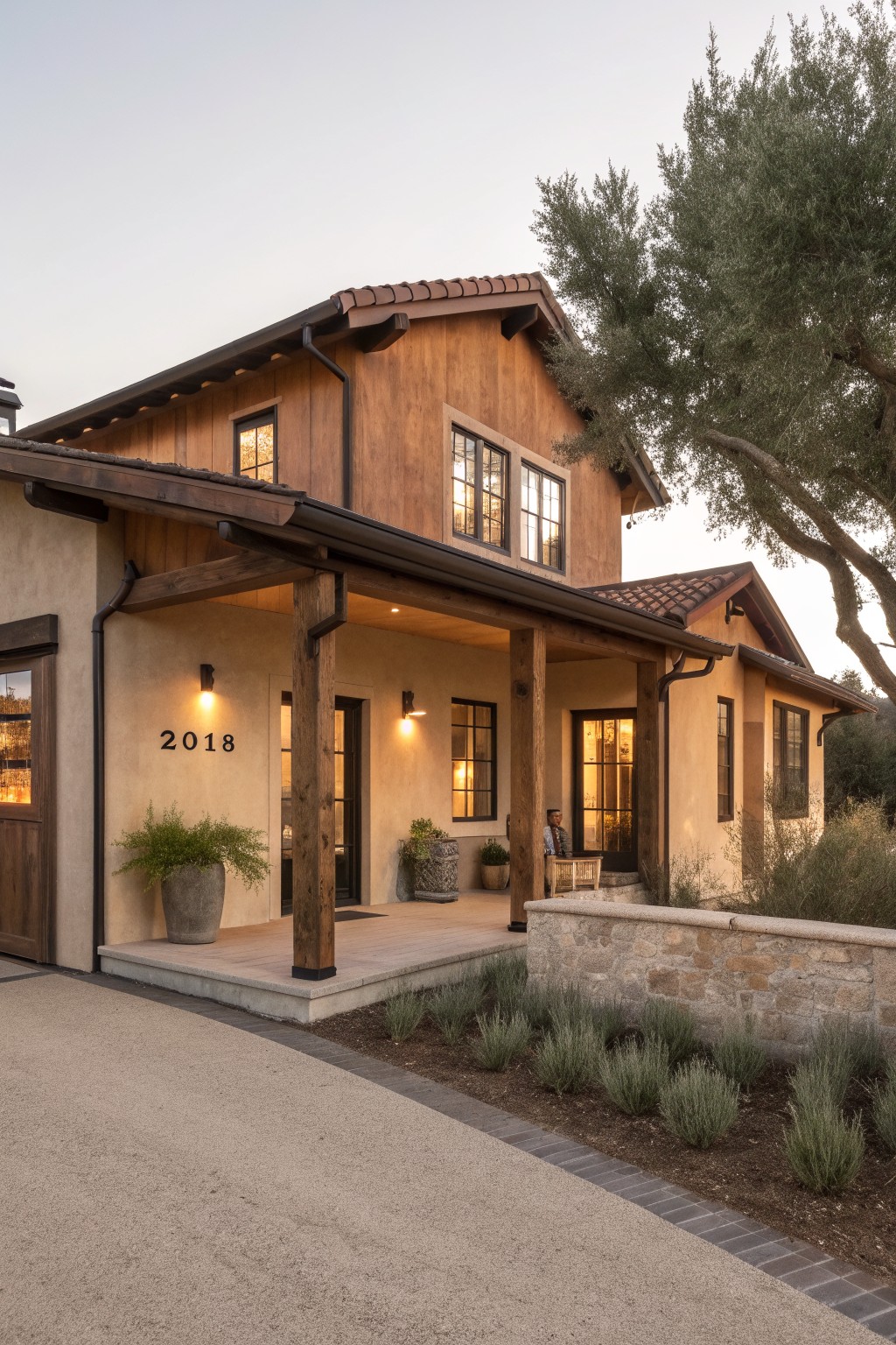 A modern barn-style house exterior with upper wood siding, lower stucco walls, a wide covered porch on thick timber posts, black-framed windows, and a stone retaining wall along the front.
