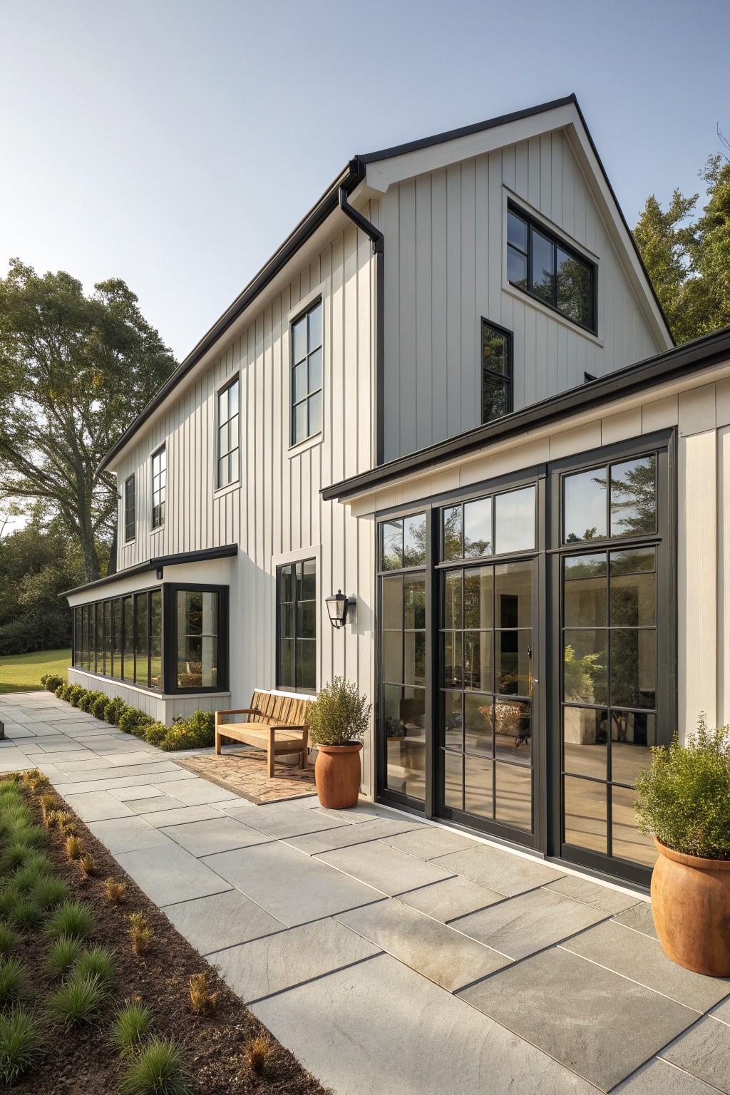 Modern barn-style house exterior with light vertical siding, black-framed windows and doors, stone patio, wooden bench, and potted plants.