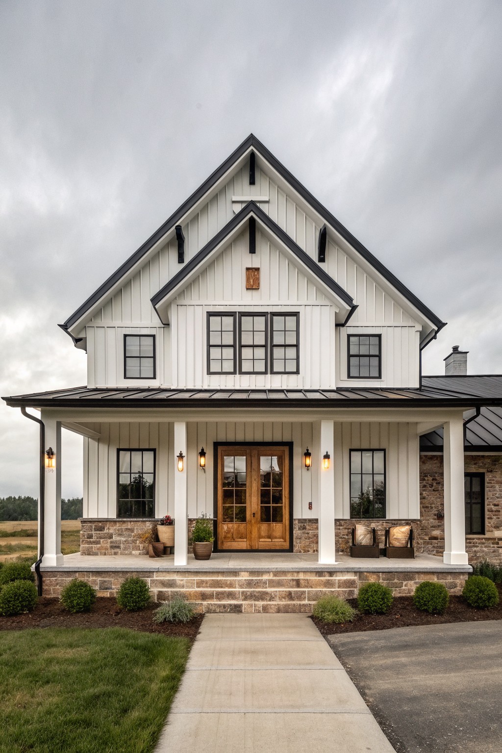 A front view of a modern two-story barn-style house with white vertical siding, black trim, a central wooden double door, stone foundation, and a covered porch.