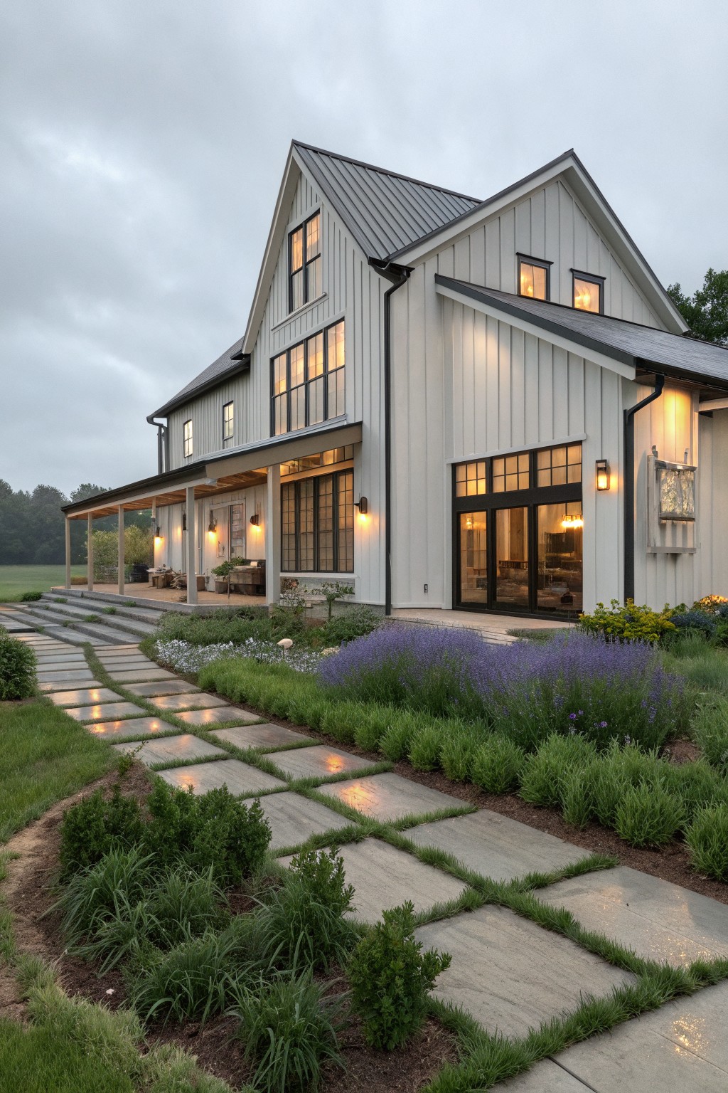 A white modern barn-style house with vertical siding, black-framed windows of various sizes, a dark metal roof, and a stone pathway lined with lavender and grasses leading to the porch.