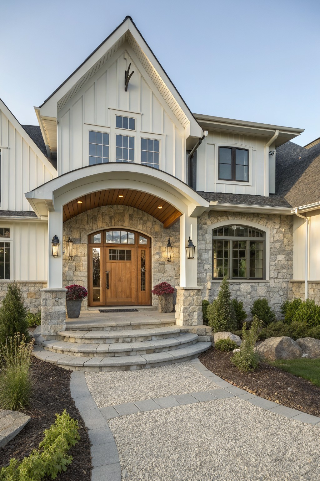 Front exterior of a two-story contemporary farmhouse with white siding, stone cladding around an arched entry, a wooden door, and a gravel driveway with stone steps.