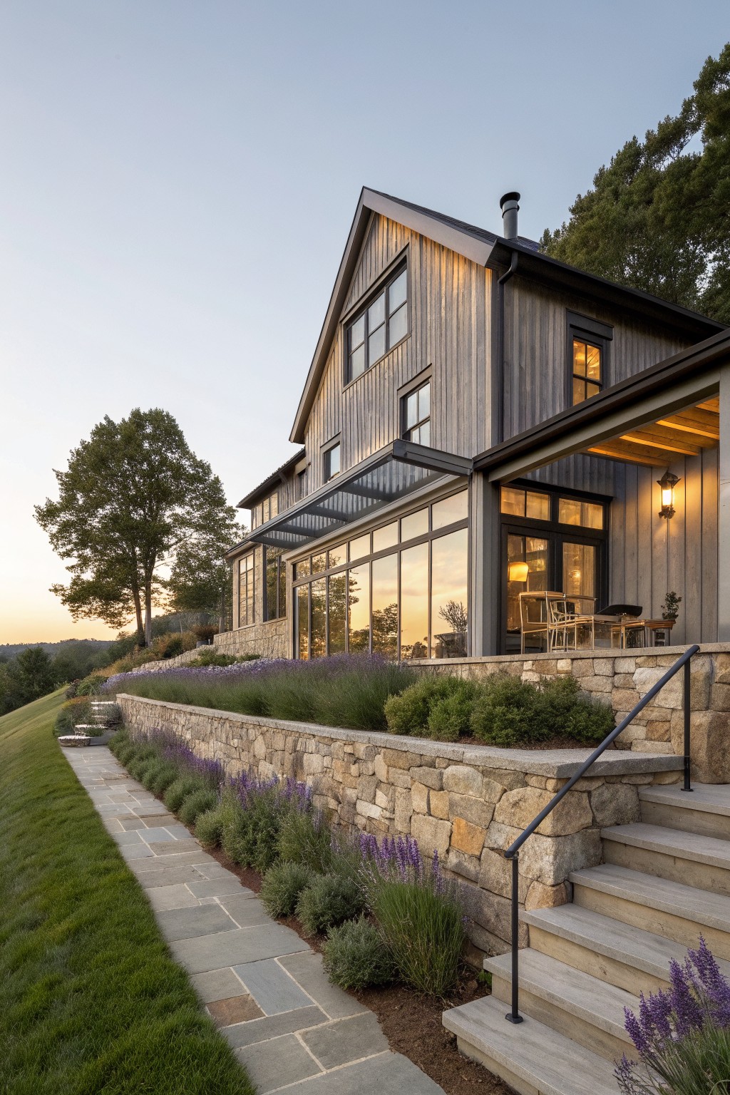 A contemporary farmhouse exterior featuring dark vertical wood siding, large glass windows, black trim, and a prominent stone retaining wall along a sloped site with steps and landscaping.