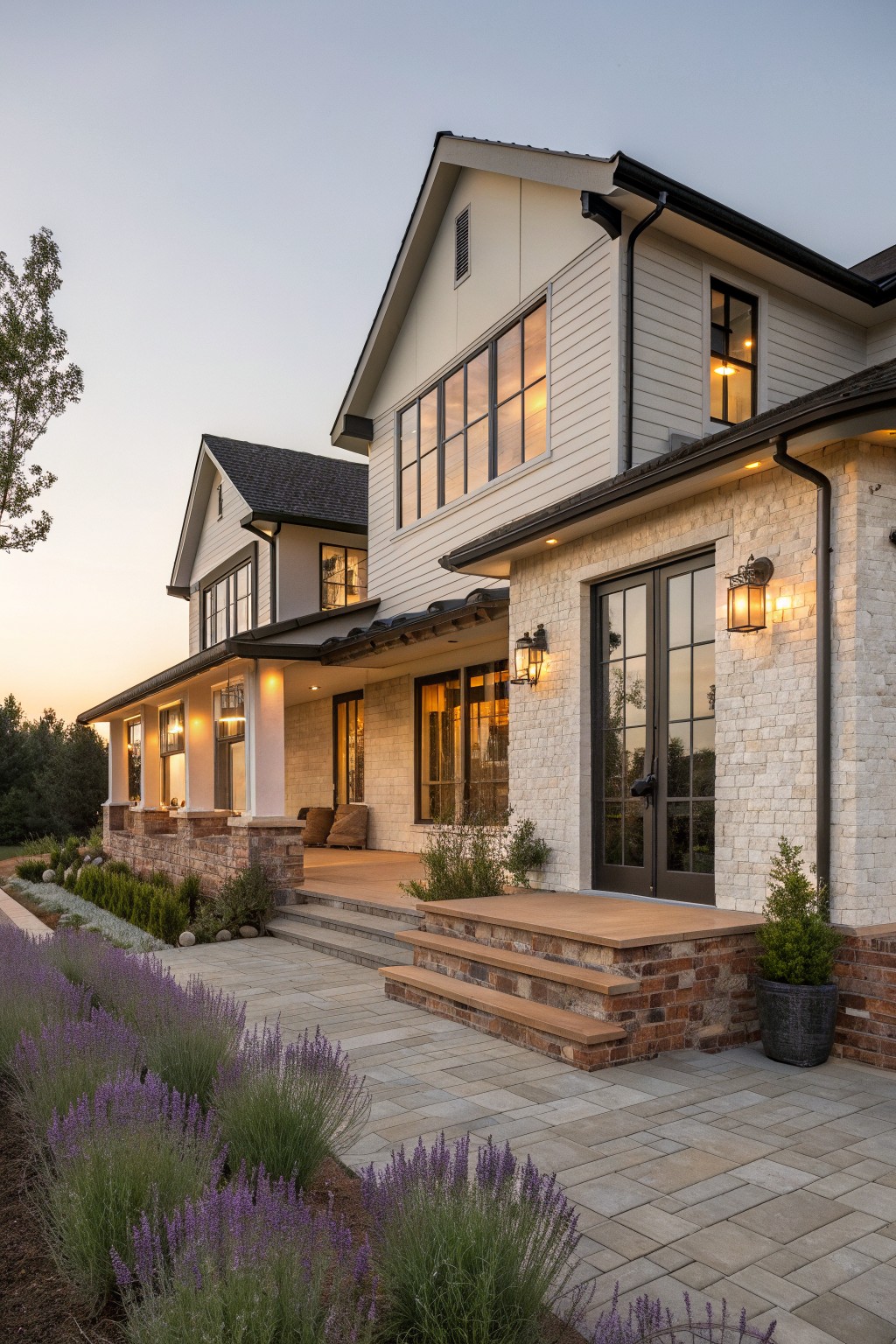 Modern two-story farmhouse with white siding, stone accents, lit windows, and lavender garden at dusk.