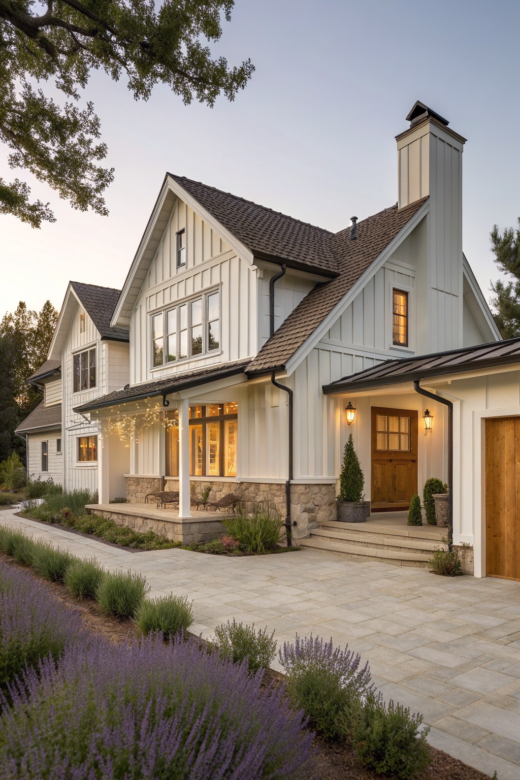 A two-story white contemporary farmhouse with vertical board and batten siding above a stone foundation, brown shingle roof, prominent chimney, wooden front door, and paved driveway with lavender in the foreground.
