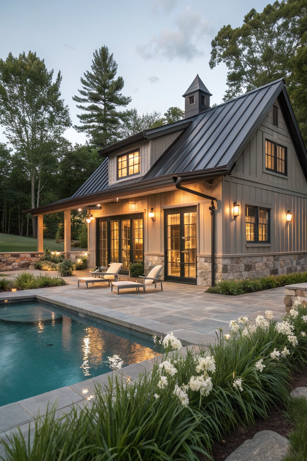 A gray rustic farmhouse with a dark metal roof, stone foundation, and a pool patio in the foreground at dusk.