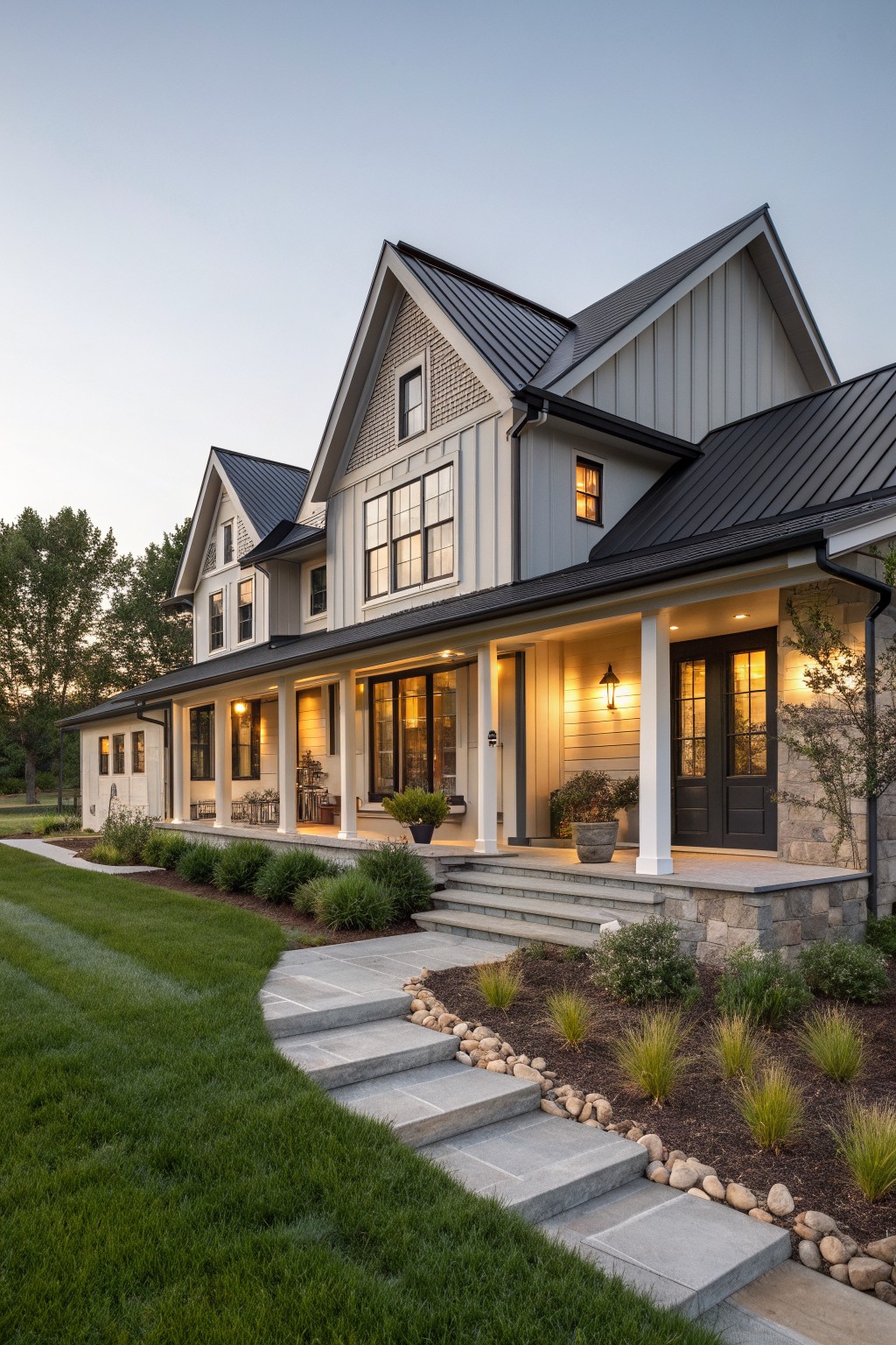 Modern gray house with black roof, white columns, lit porch, stone steps and lawn.