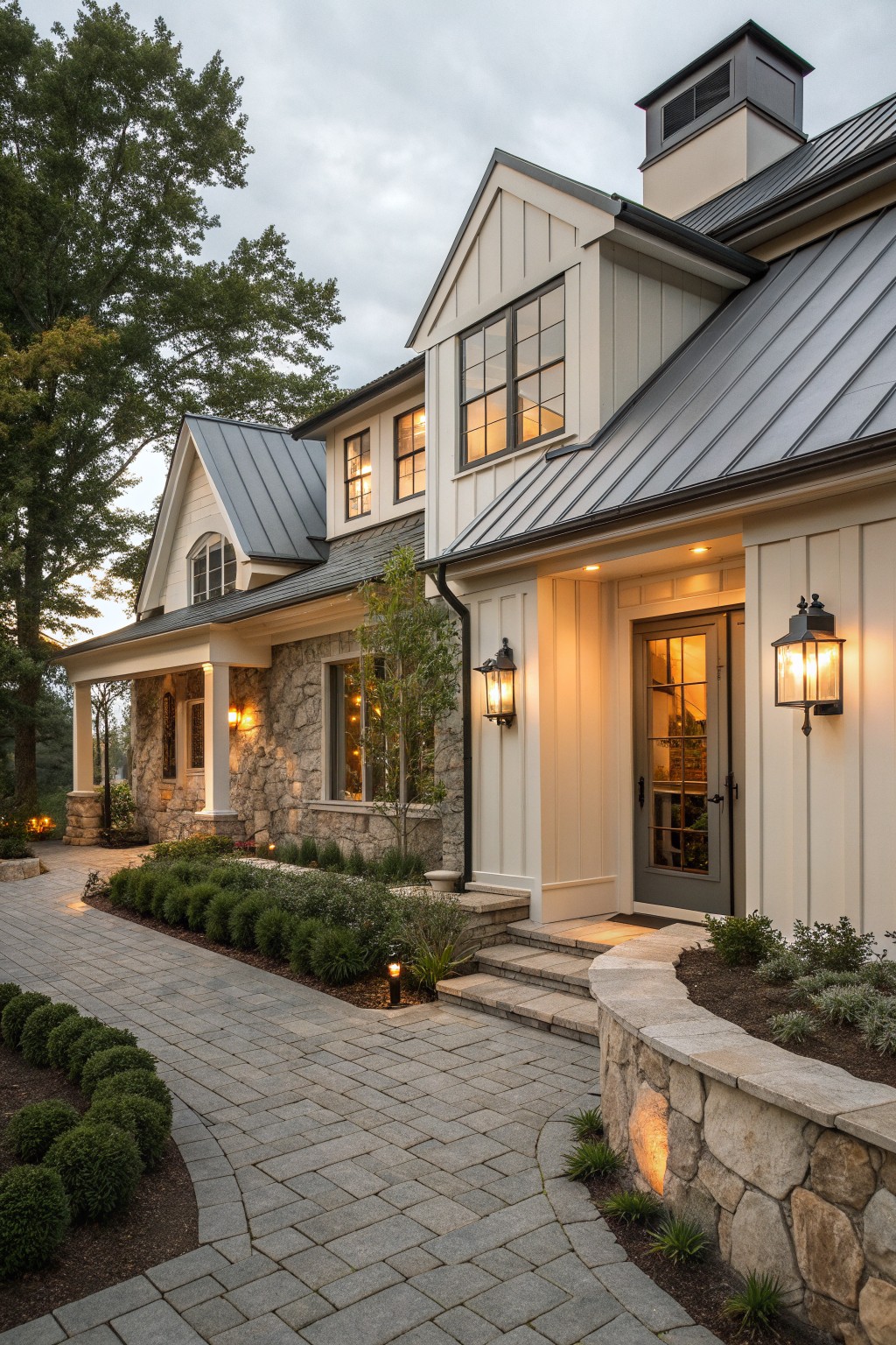 A front view of a two-story farmhouse with stone walls on the lower level, white board and batten siding above, a gray metal roof, and a central entry door with stone steps and wall lanterns.