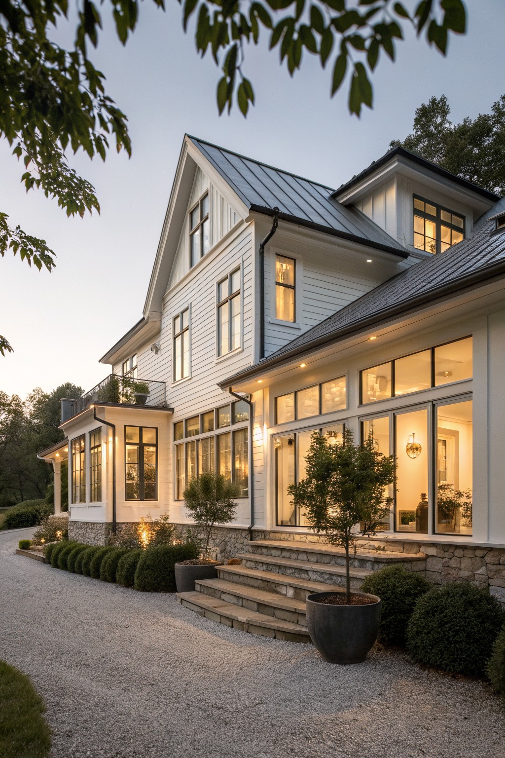 A white two-story farmhouse at dusk with a long row of large multi-pane windows across the main level above a stone foundation, a gravel driveway in front, and potted plants near the steps.