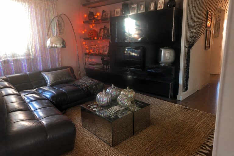 18 2000s Halloween Decor Ideas That Are So Y2K It’s Scary A living room with a black leather sectional, mirrored coffee table holding two iridescent pumpkins, chrome floor lamp, and black built-in shelves with string lights.