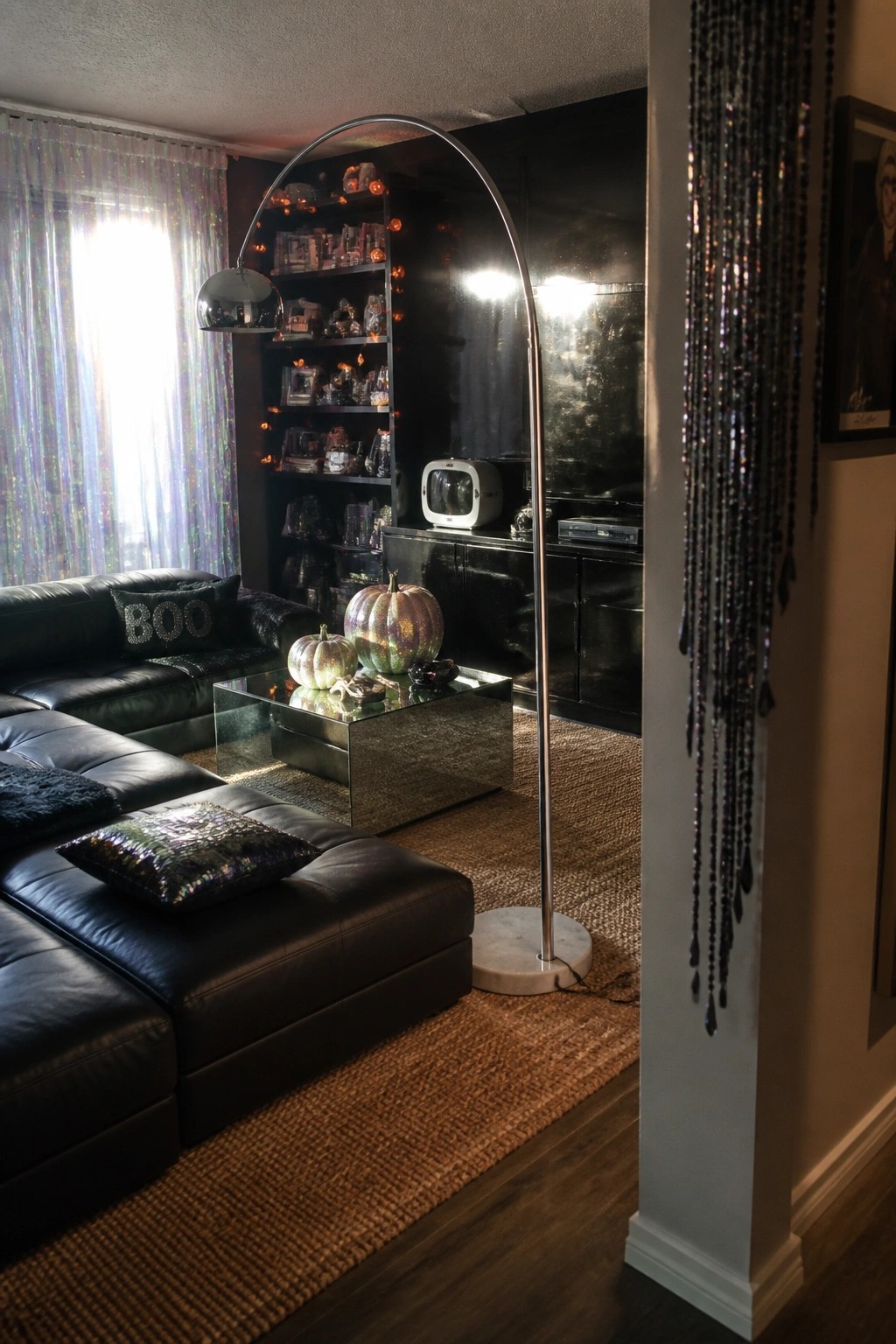 A living room with a black leather sectional, mirrored coffee table holding two iridescent pumpkins, chrome floor lamp, and black built-in shelves with string lights.