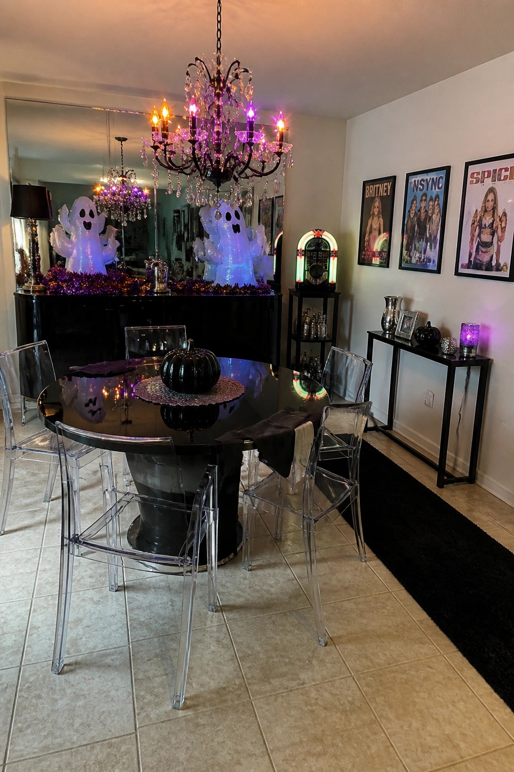 A dining room with a round black table, transparent chairs, inflatable ghosts, purple string lights, and three framed 2000s pop star posters on the wall.