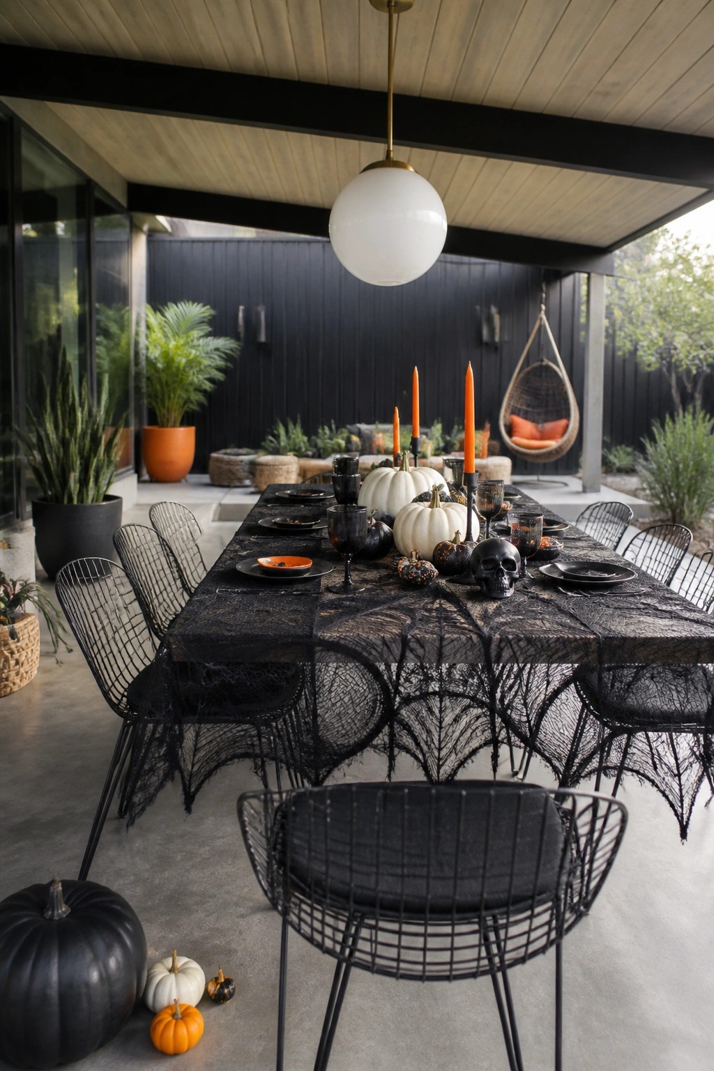 Outdoor patio dining table covered in black spiderweb lace with orange taper candles, black glassware, a skull, and mixed black and white pumpkins.
