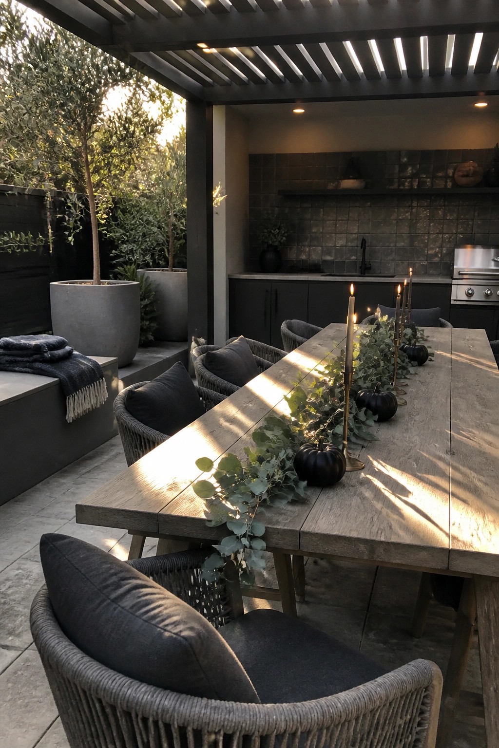 Outdoor wooden dining table with black pumpkins, eucalyptus garland, and lit taper candles as a centerpiece, surrounded by woven chairs under a pergola.
