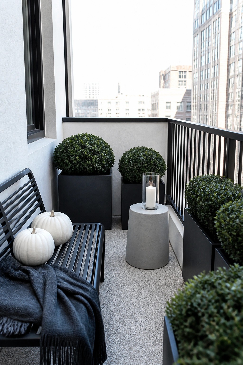 A city balcony shows a black bench with two white pumpkins and a dark throw, black rectangular planters holding round shrubs, and a lit candle on a round concrete stool.