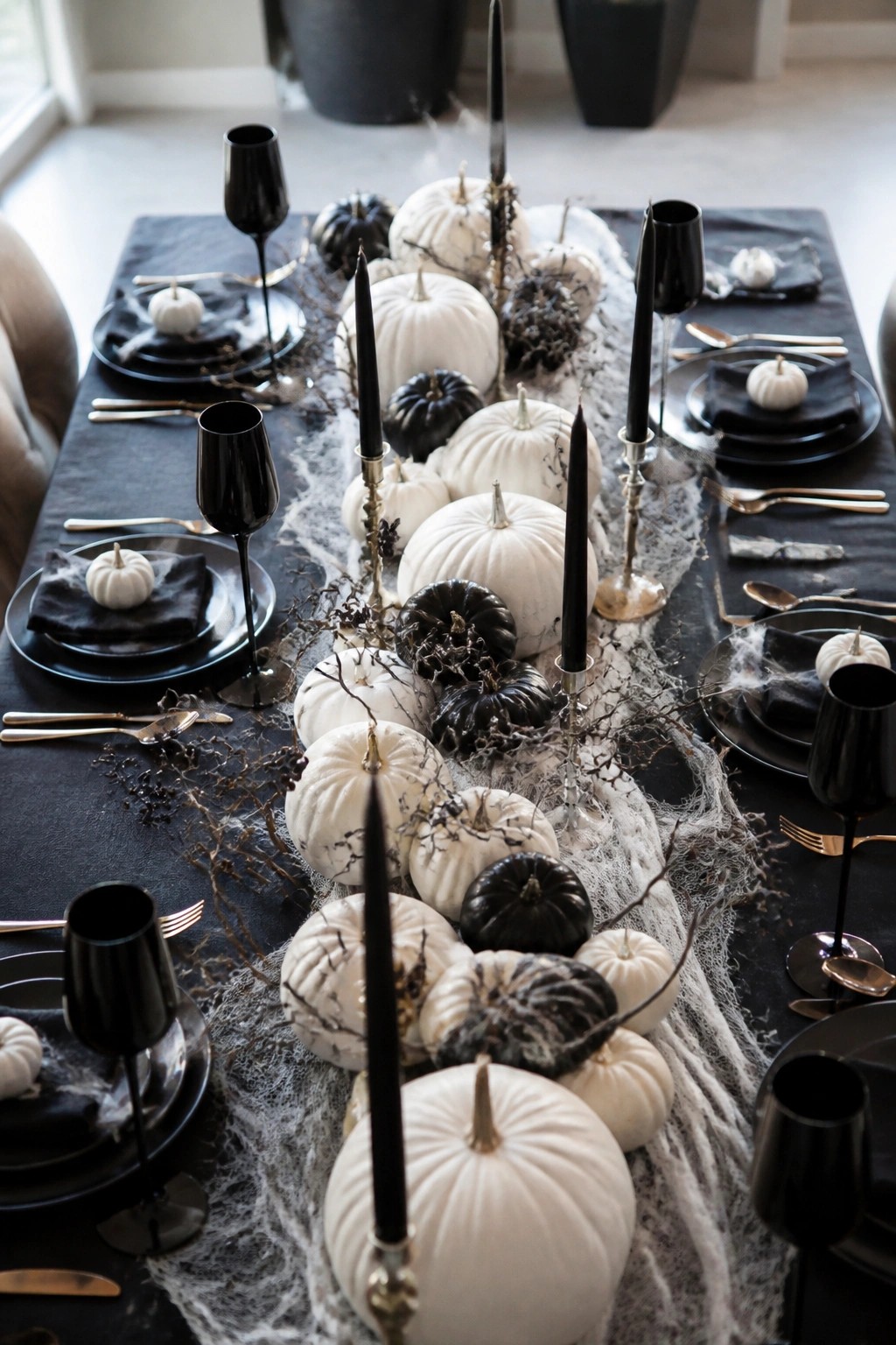 A long dining table with a black tablecloth features a centerpiece of white and black pumpkins, tall black candles, and spiderwebs running down the middle.