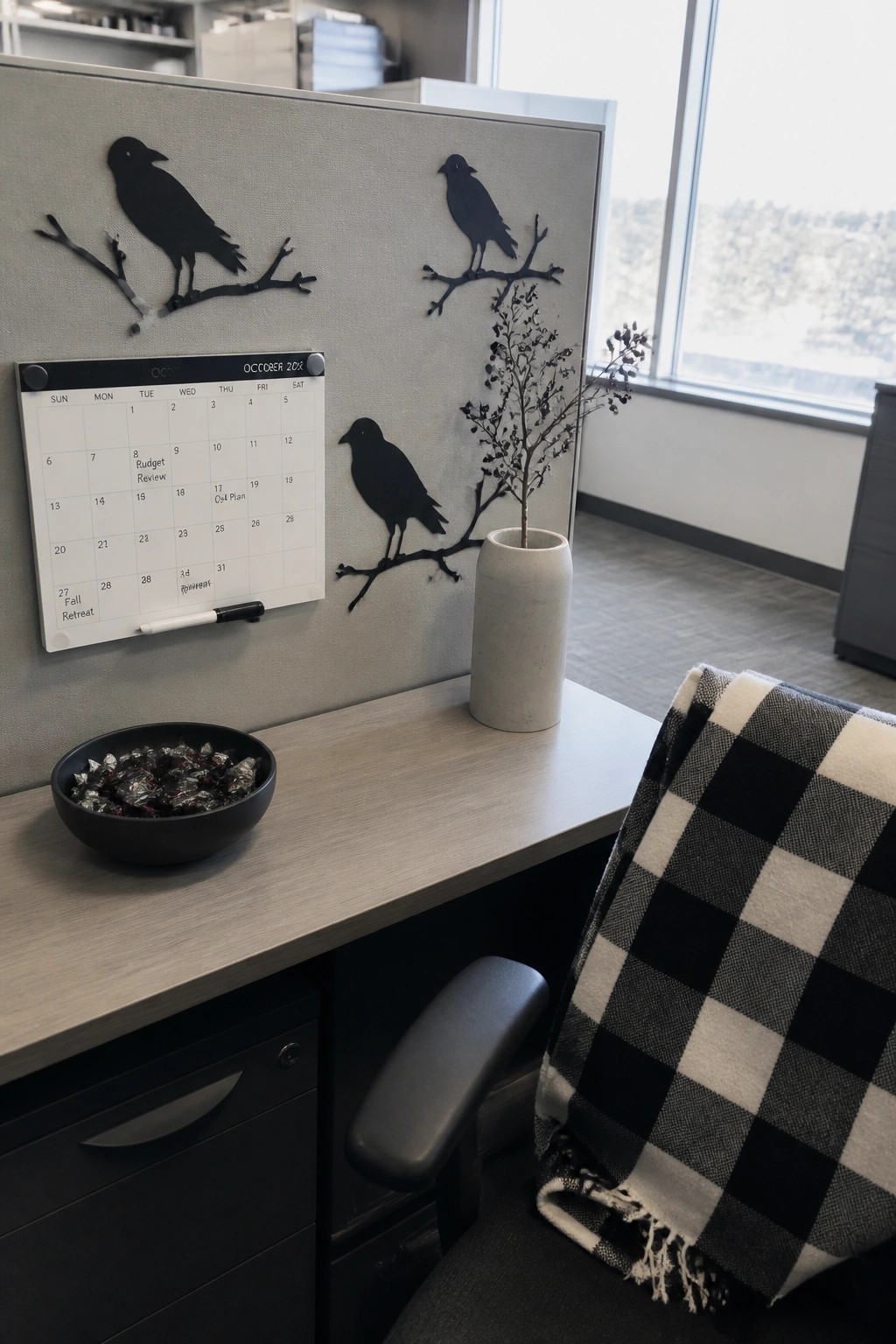 An office cubicle wall with black crow silhouettes, an October calendar, a bowl of wrapped candy, and a vase of dried branches on the desk.