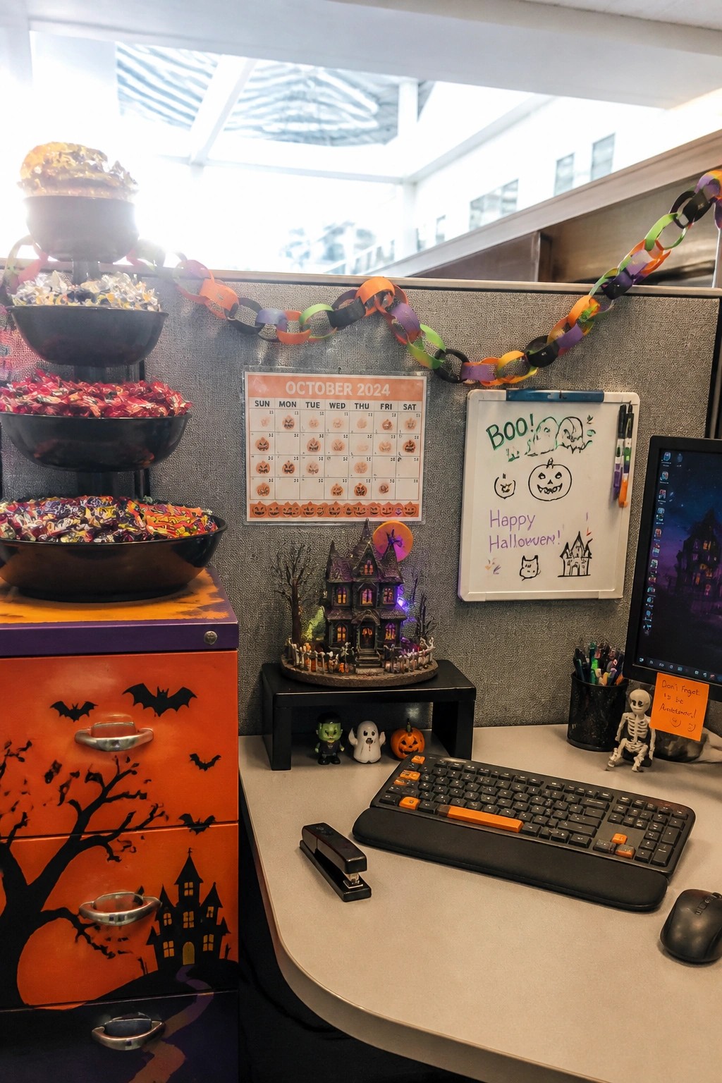 An office cubicle with a Halloween-decorated orange and purple filing cabinet holding stacked candy bowls, a calendar on the wall, a whiteboard with drawings, paper chain garlands, and small figurines on the desk.