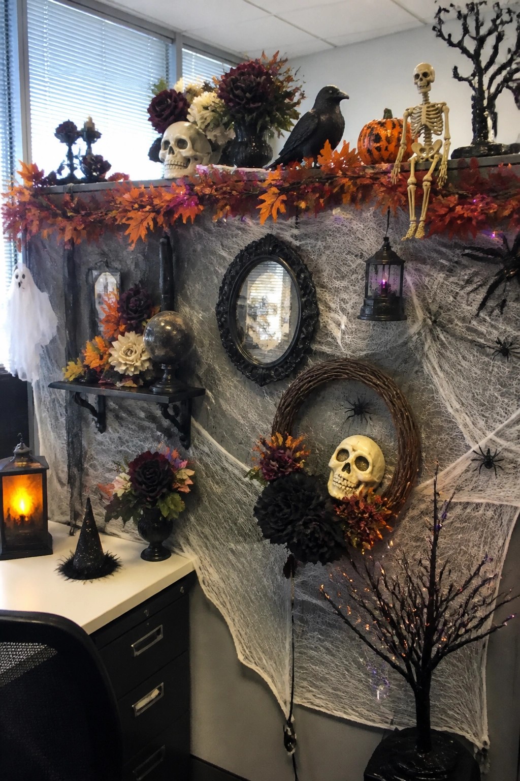 Office cubicle walls covered in white spiderwebs with skulls, skeletons, crows, and orange garlands arranged on top.