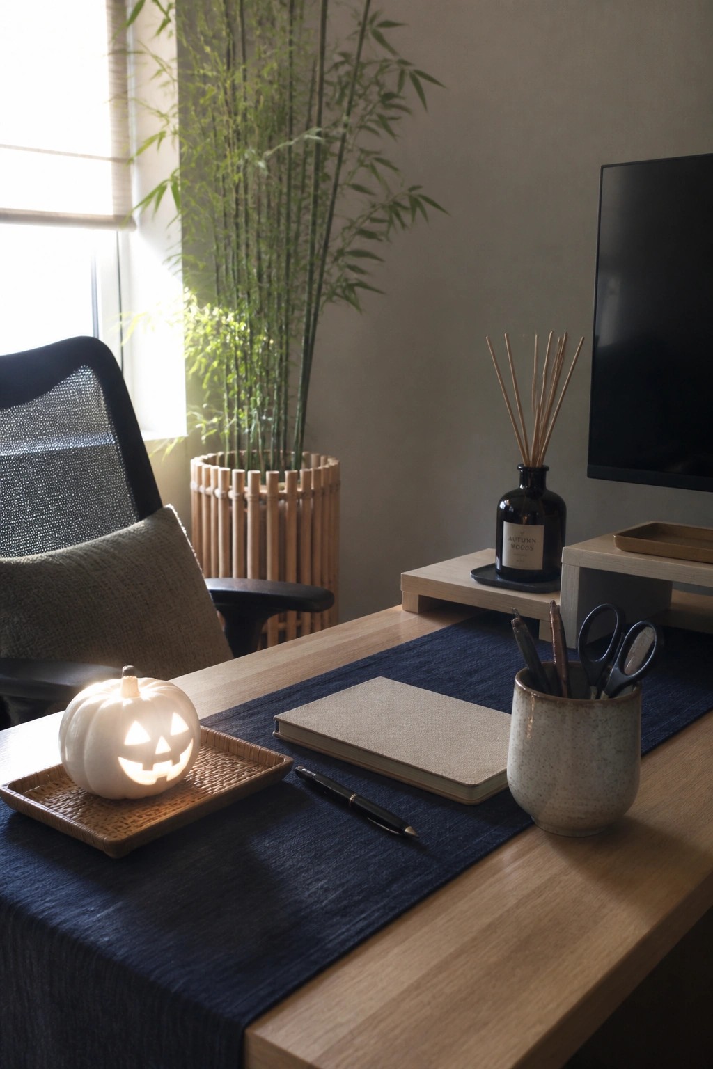 A wooden desk with a lit white jack-o'-lantern pumpkin on a woven tray, a notebook, pen, and reed diffuser on a blue runner.