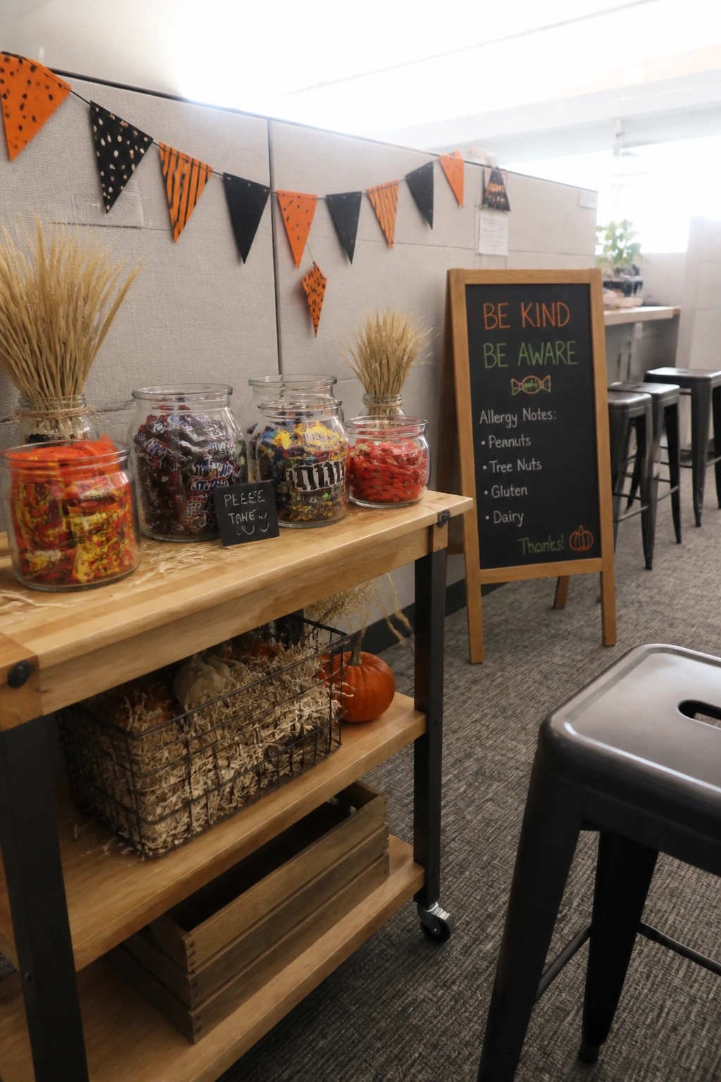 A wooden table in an office holds glass jars of assorted candy, a small chalkboard sign, wheat bundles, and a pumpkin, with a larger chalkboard easel nearby listing allergy notes.
