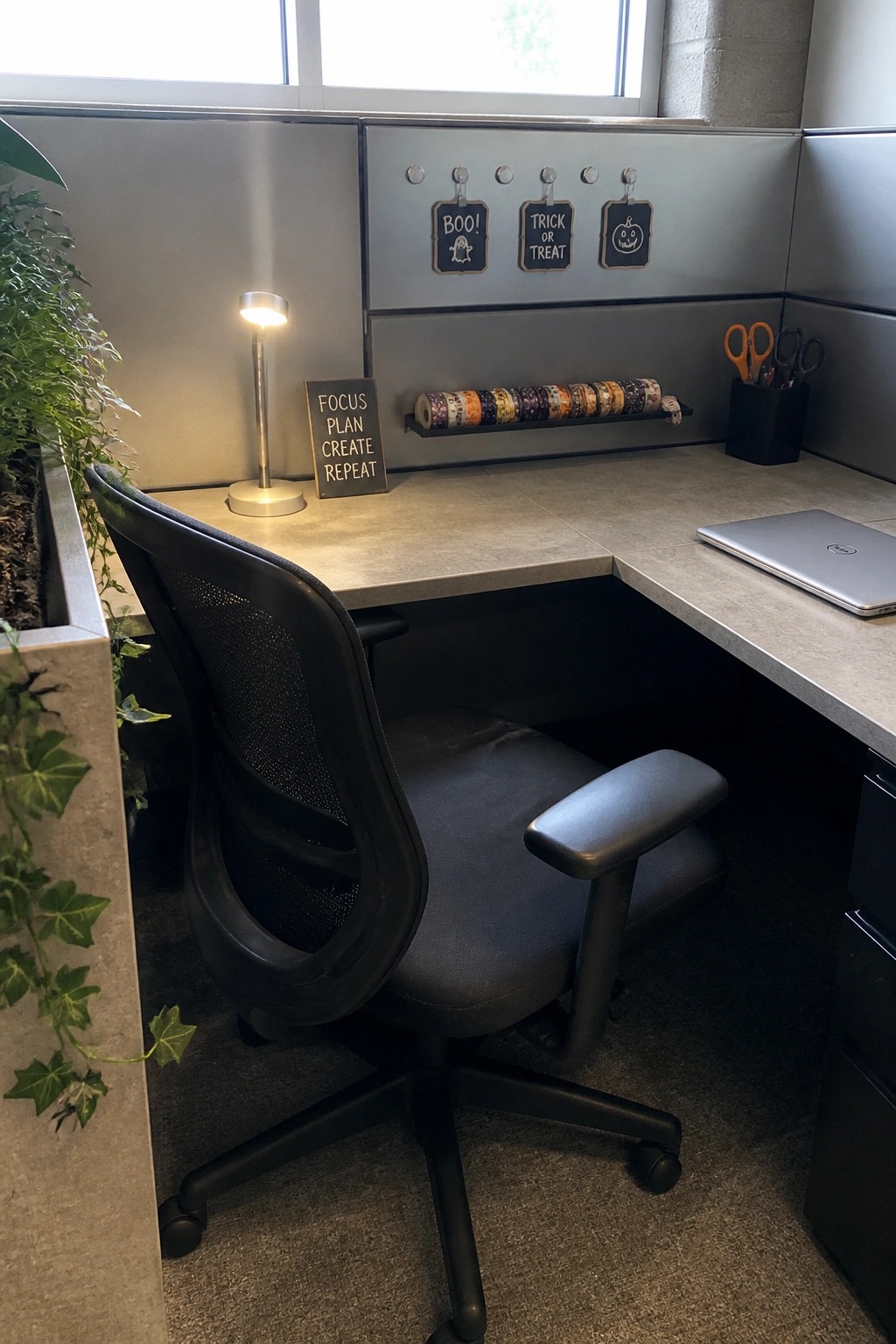 A cubicle workspace with a black office chair, desk lamp, motivational sign, roll of Halloween washi tape, and three small chalkboard signs on the wall.