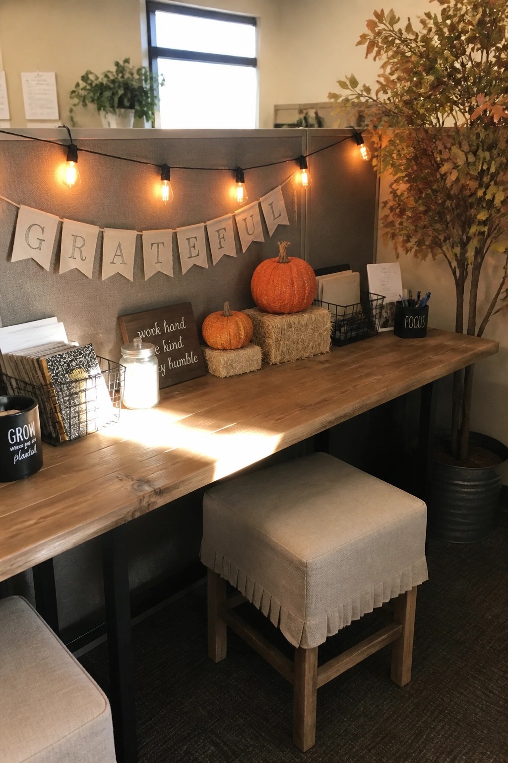 An office cubicle desk with string lights, a grateful banner, pumpkins, hay bales, and office supplies arranged on a wooden surface.