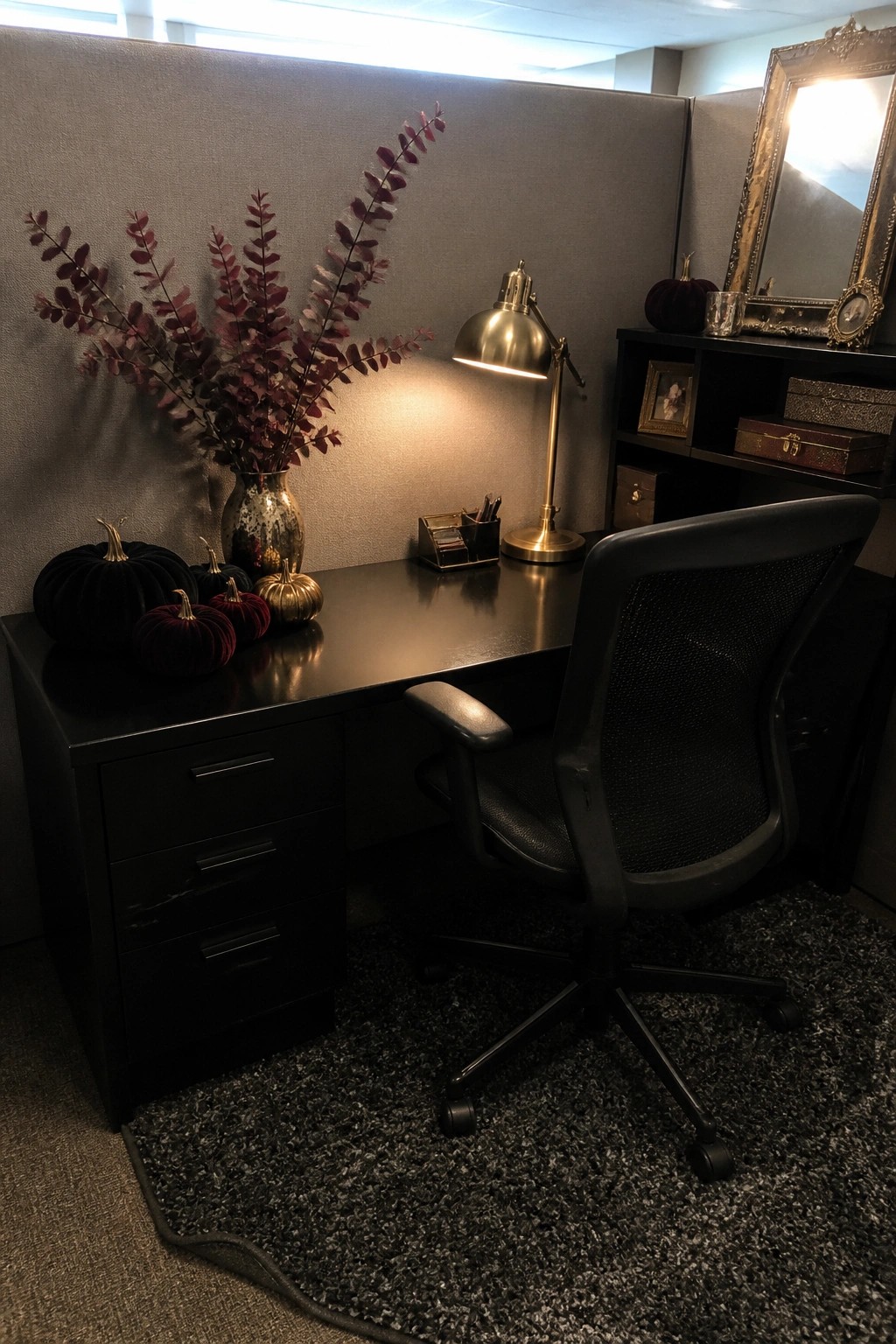 A black office desk with several velvet pumpkins in black, red, and gold tones plus dark red foliage in a vase, lit by a brass desk lamp with a black office chair pulled up to it.