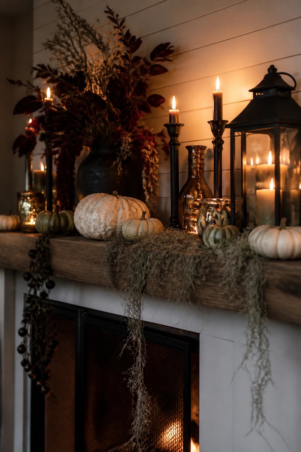 Wooden mantel decorated with black candlesticks, a black lantern, white and green pumpkins, and dried foliage above a white fireplace.