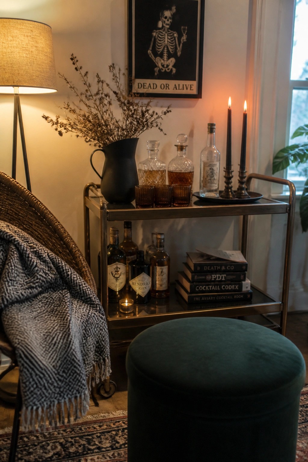 A metal bar cart holds liquor bottles, amber glasses, black taper candles, and a black pitcher with dried branches, with a skeleton poster hanging above it.