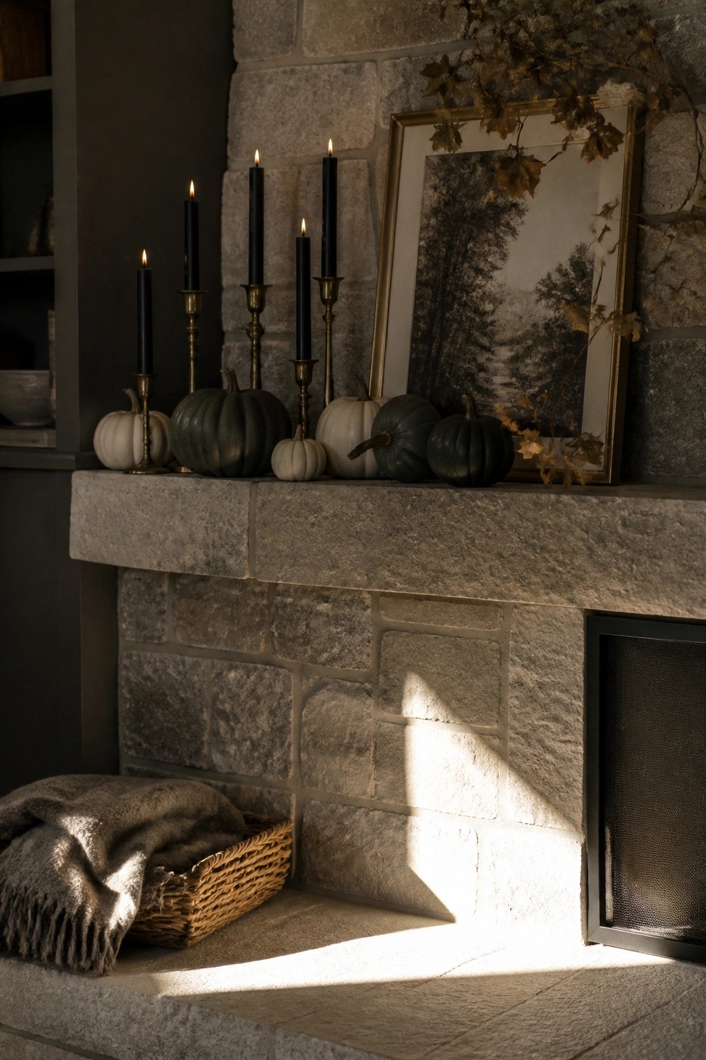 A stone fireplace mantel displays five black taper candles in brass holders, several small pumpkins in white and dark shades, a framed black and white landscape photo, and dried leaves draped along the right side, with a wicker basket holding a folded blanket on the hearth below.