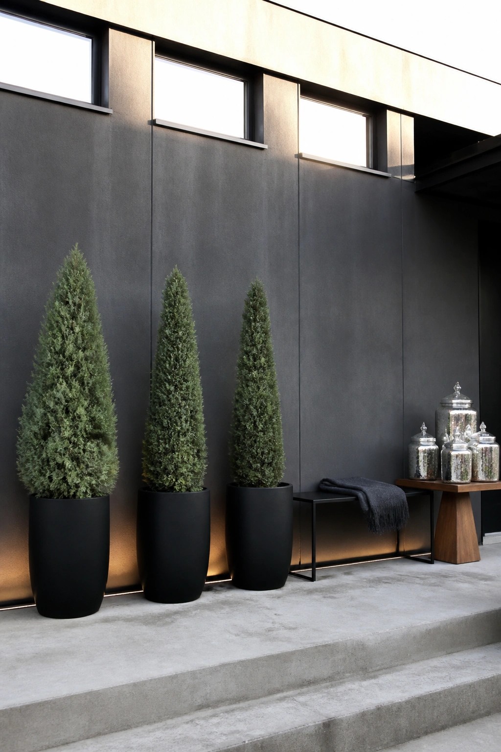 Three tall evergreen trees in large black pots stand in a row against a dark exterior wall with small windows, next to a bench and wooden table holding silver jars.