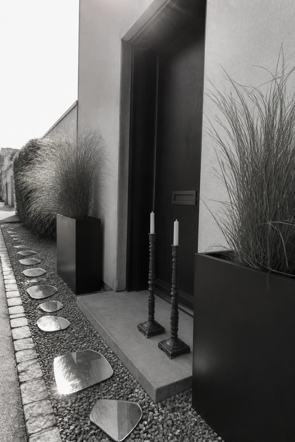 Modern entrance with dark door, candlesticks, potted grasses, and reflective stepping stones on gravel