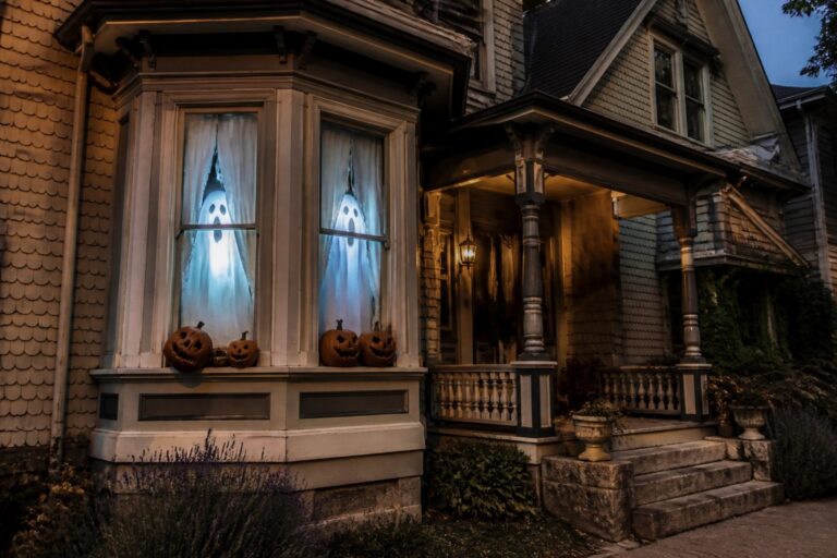 18 Scary Halloween Window Ideas That’ll Shock the Neighborhood A gray house at dusk with three illuminated ghost figures visible through a bay window and carved pumpkins placed along the windowsill.