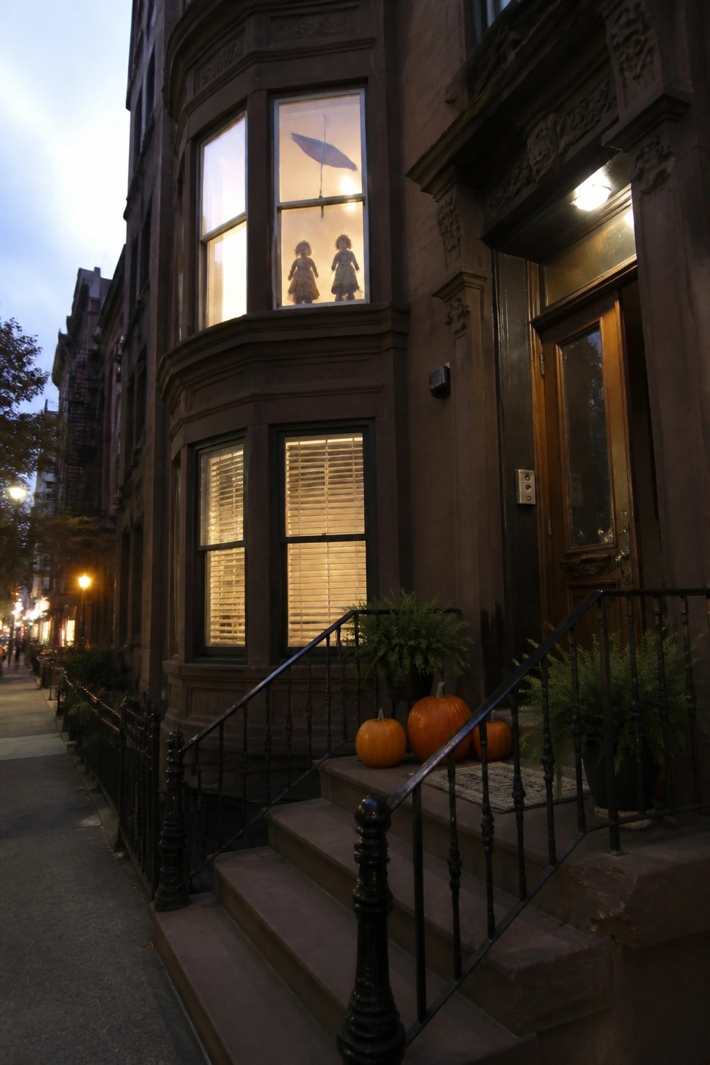 Two dolls hanging inside an upper window of a brownstone building at dusk, with pumpkins placed on the front steps.