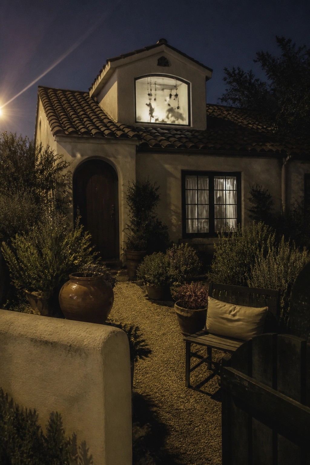 A nighttime view of a stucco house exterior showing a lit gable window with hanging decorations, a gravel path, and potted plants along the front.