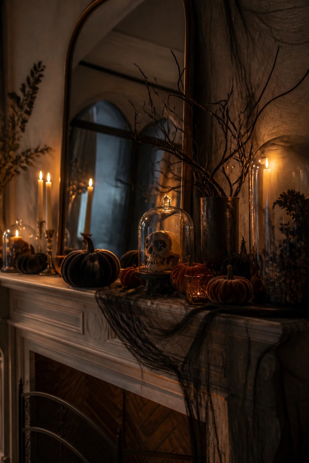 White fireplace mantel decorated with black spiderwebs, velvet pumpkins, lit candles, bare branches, and a skull under a glass cloche.
