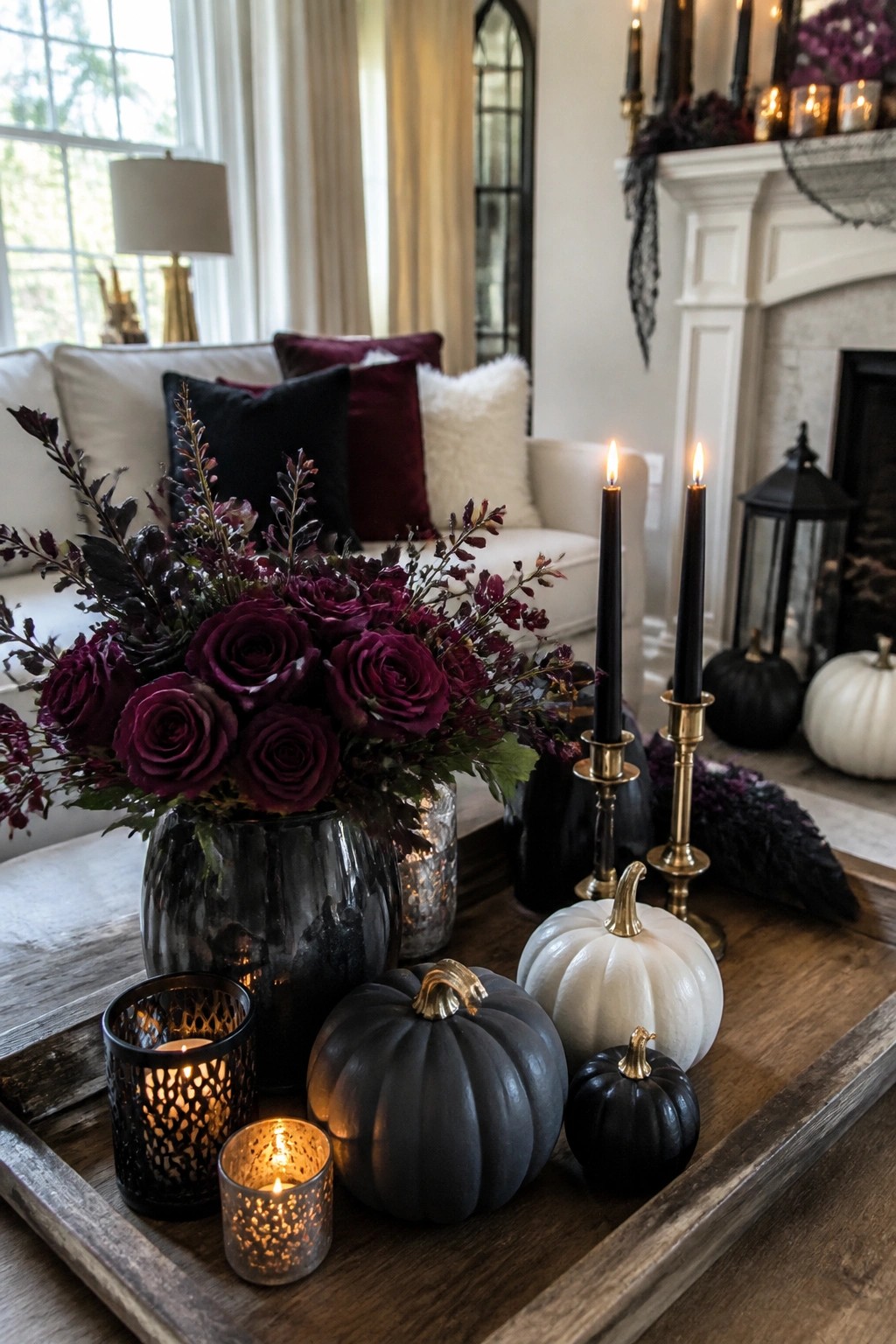 Wooden tray on a coffee table holding black and white pumpkins, black taper candles in gold holders, and a dark floral arrangement with purple roses.