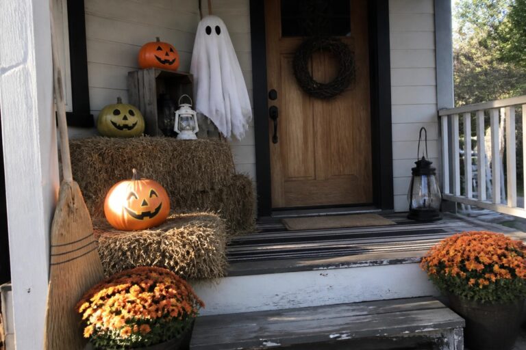 20 DIY Outdoor Halloween Decor Ideas That Won’t Break the Bank A wooden front porch decorated with hay bales holding carved pumpkins, a sheet ghost on a stake, two lanterns on a crate, a wreath on the door, and potted orange mums.