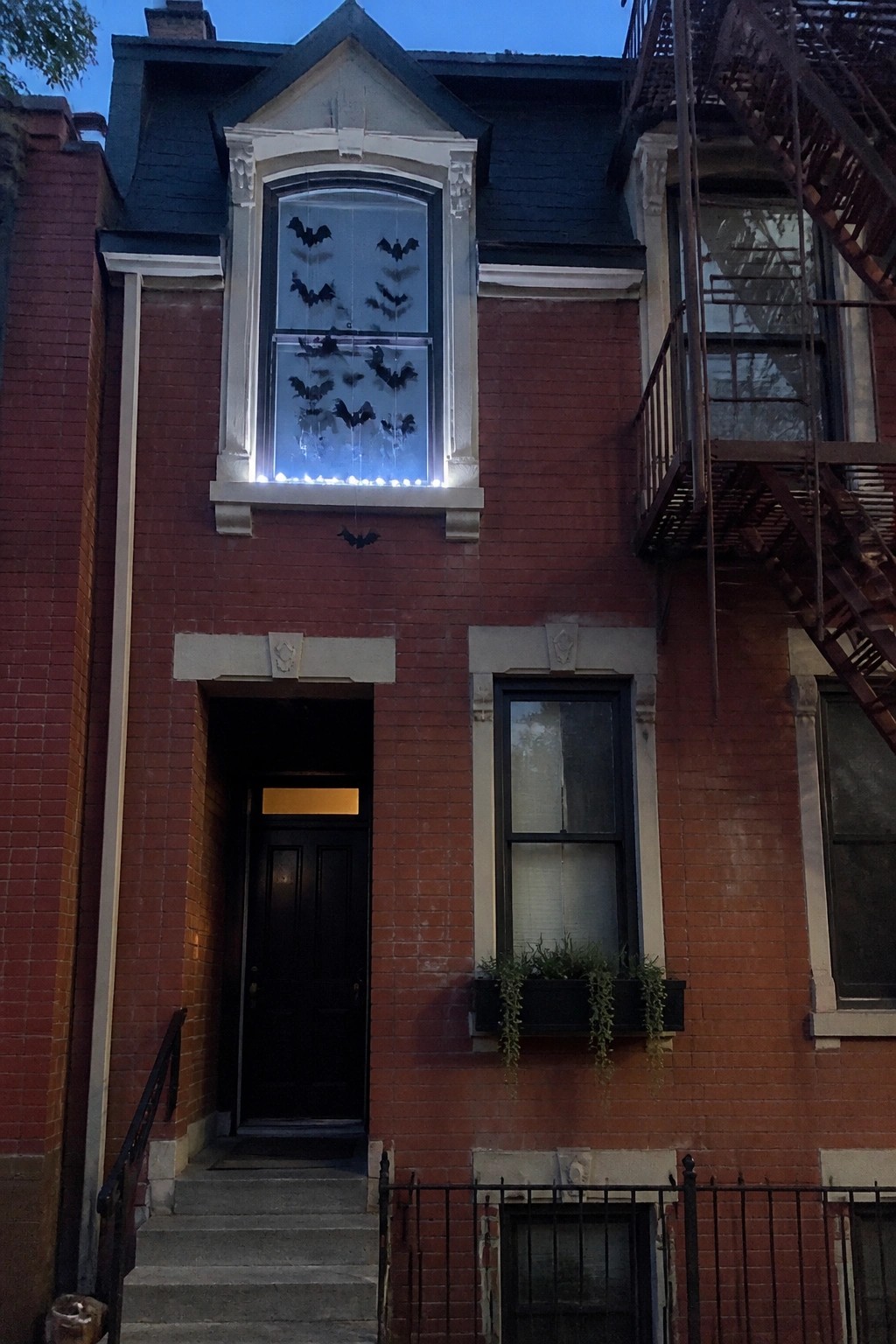 A red brick house facade at dusk showing a large upper window filled with black bat cutouts and illuminated by string lights along the sill.