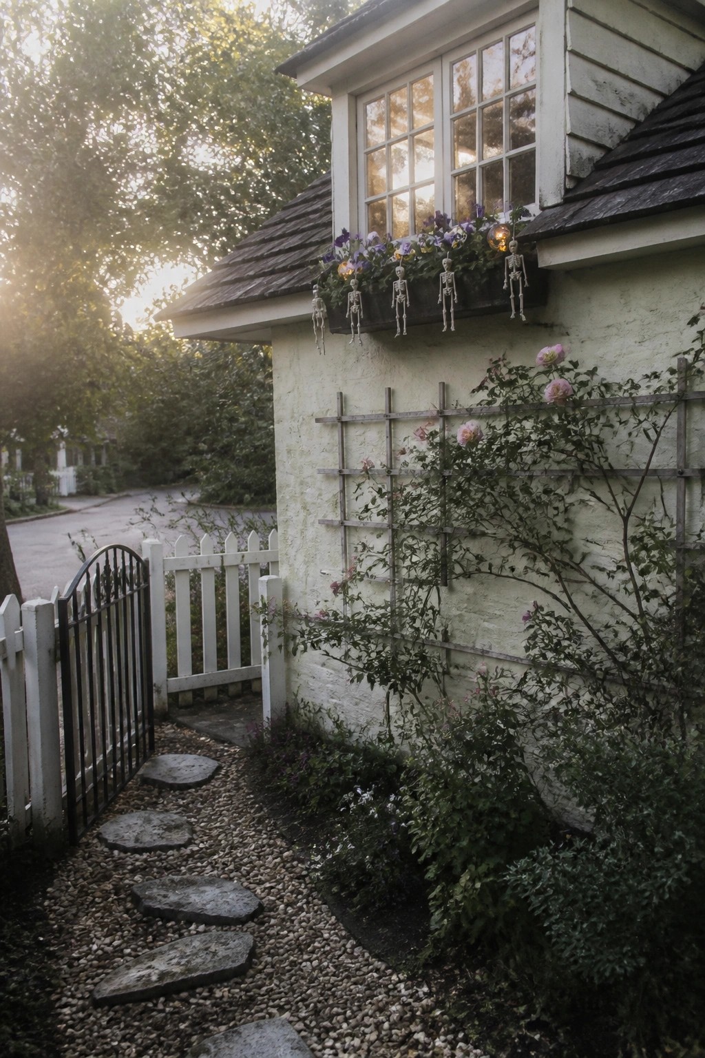 A house exterior with small white skeleton figures hanging from the flower box under a multi-paned window, next to climbing roses and a gravel path.
