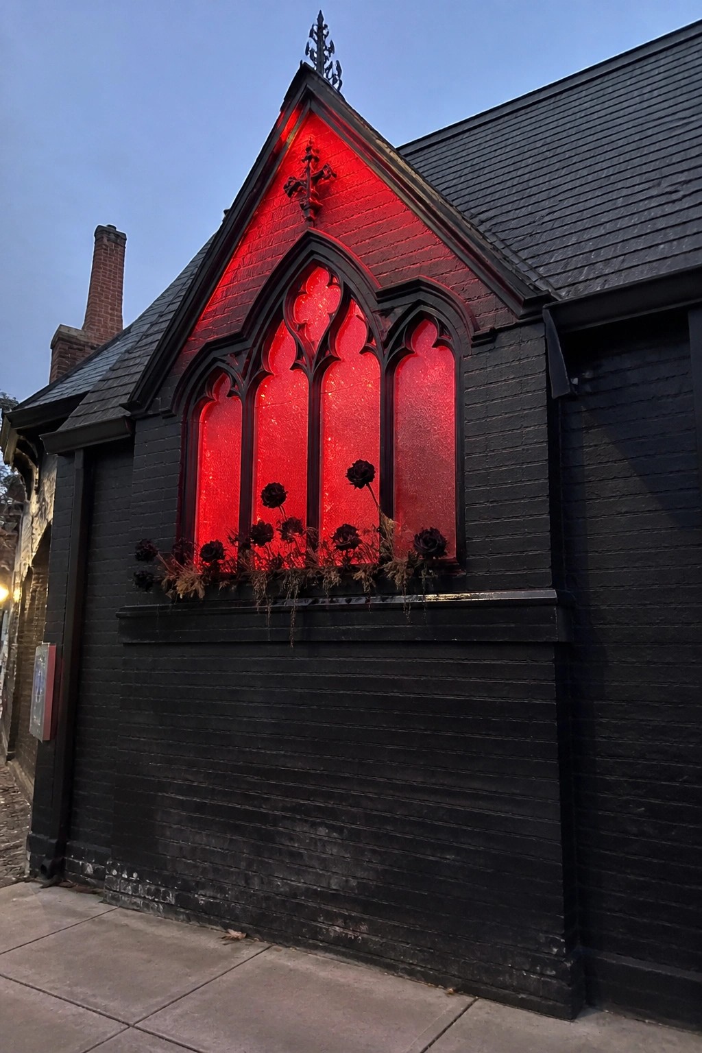 A black house with a large multi-pane Gothic window glowing bright red from within, with dark flowers along the sill.