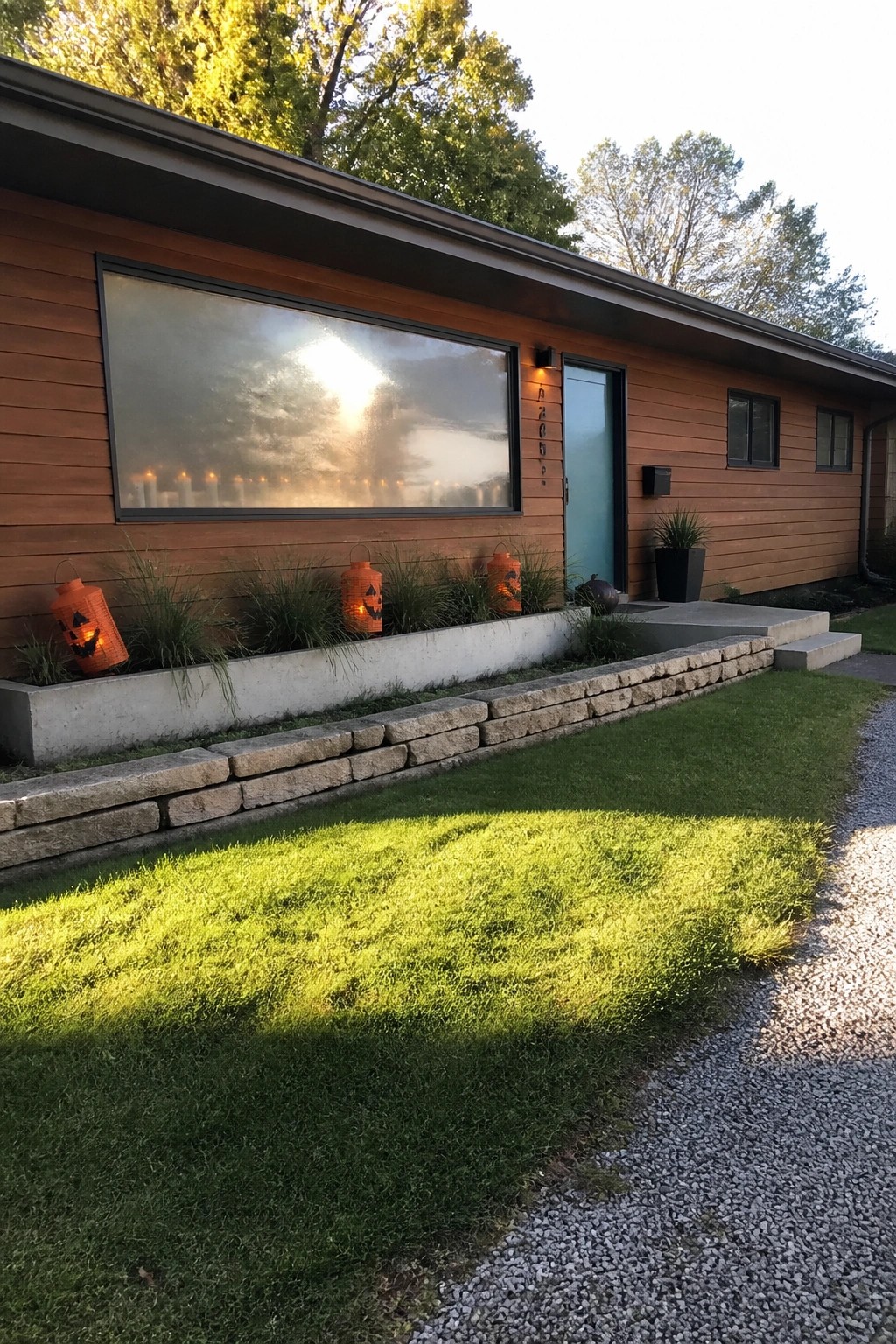 A modern wooden house with a large front window filled with lit candles and several orange jack-o-lantern lanterns placed in the garden bed below.