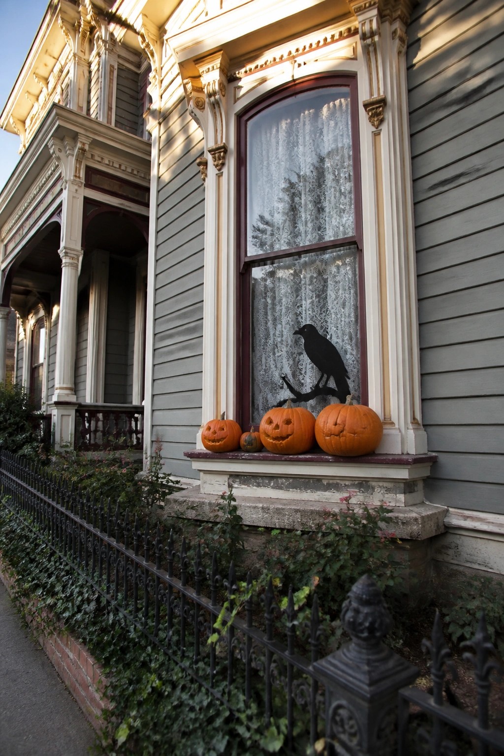 A gray Victorian house exterior with a large window displaying a black bird silhouette on white lace curtains and three carved pumpkins on the sill.