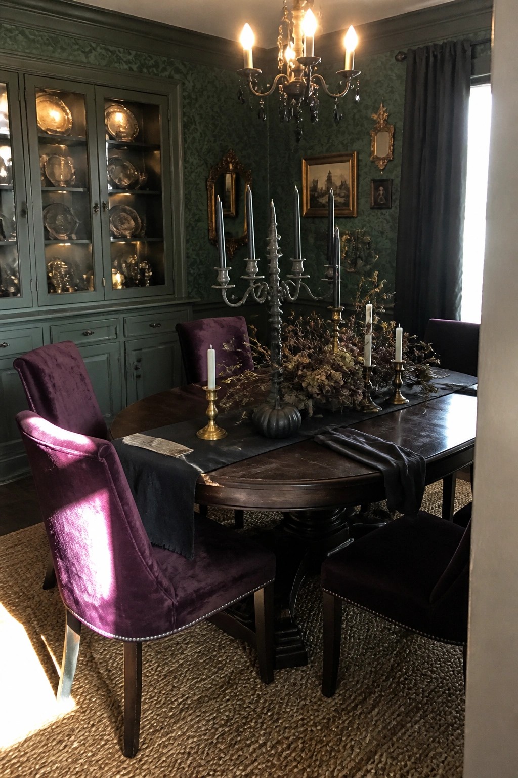 A formal dining room with dark green walls, purple velvet chairs, a round wooden table set with black candles, dried flowers, and a black table runner.