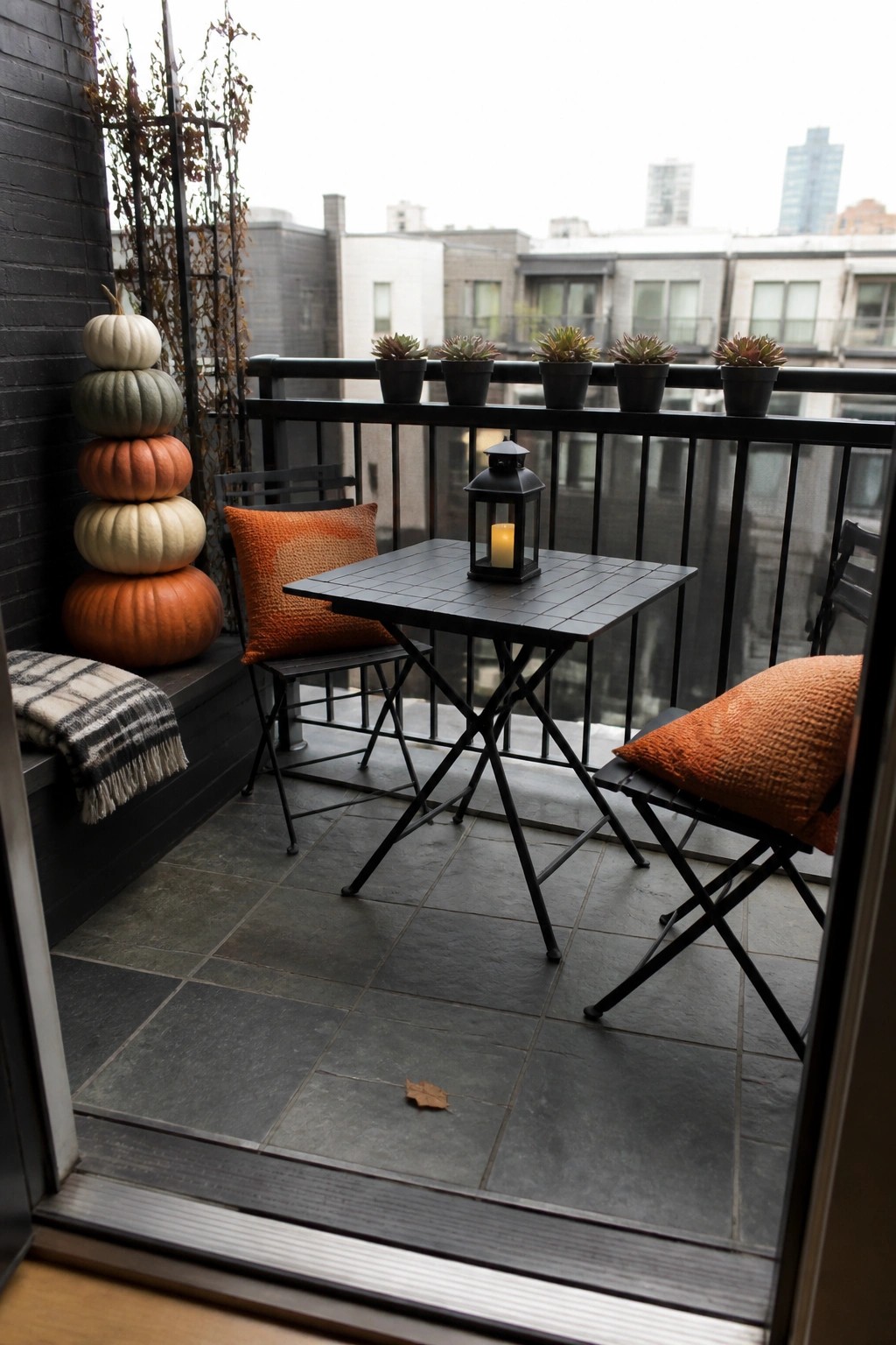 A balcony with a black folding table and chairs, orange cushions, a lit lantern on the table, potted succulents along the railing, and a tall stack of pumpkins on the left side.