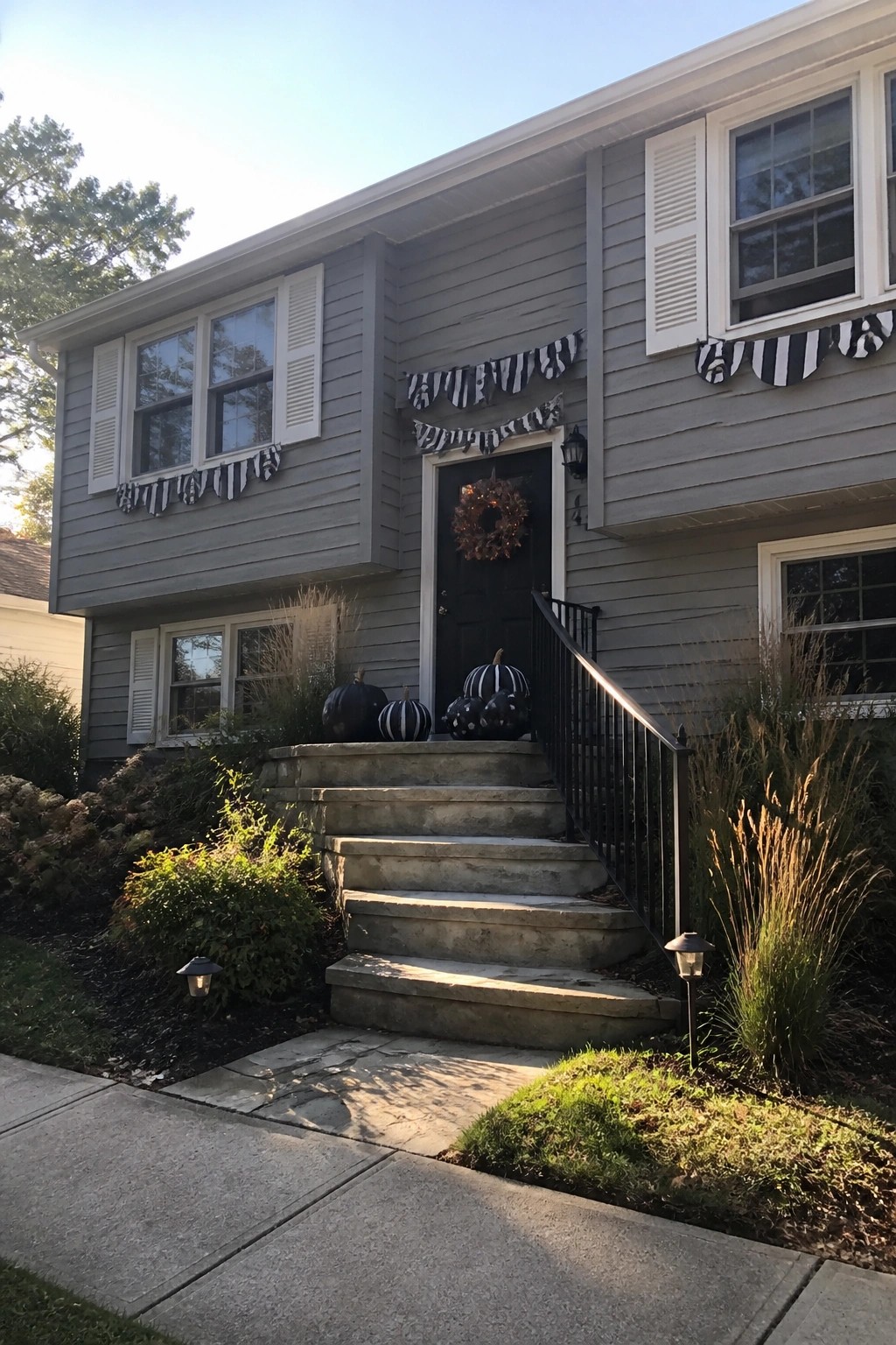 Front view of a gray two-story house with black door, black and white striped bunting across the facade, black pumpkins on the steps, and a wreath on the door.