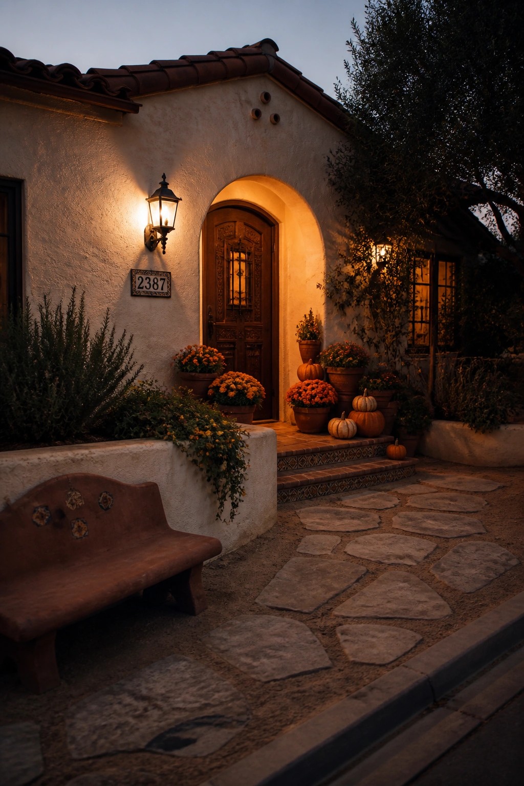 A stucco house at dusk with an arched wooden door, warm lantern light, stone pathway, potted orange flowers, and several pumpkins arranged on the entry steps.