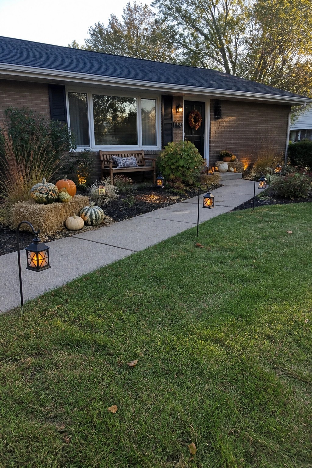 A brick house with a concrete front walkway lined by black lanterns, pumpkins on hay bales, and autumn plants in the garden beds.