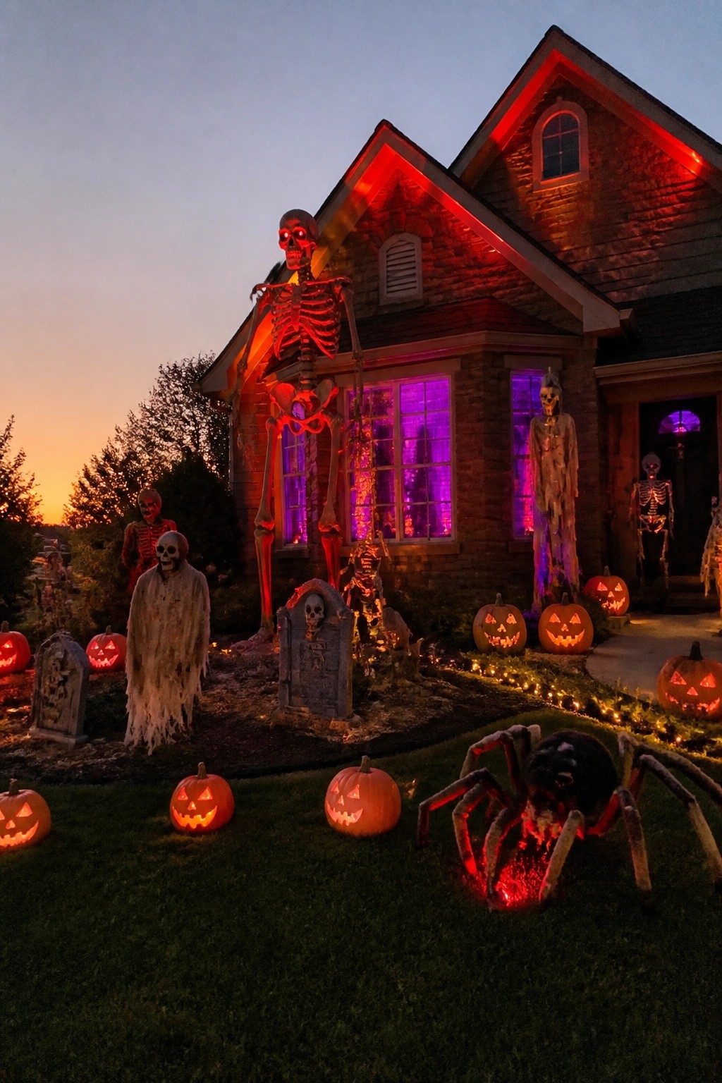 Large house exterior decorated with a giant red-lit skeleton on the front, smaller skeletons, gravestones, carved pumpkins, and a large spider prop in the yard.
