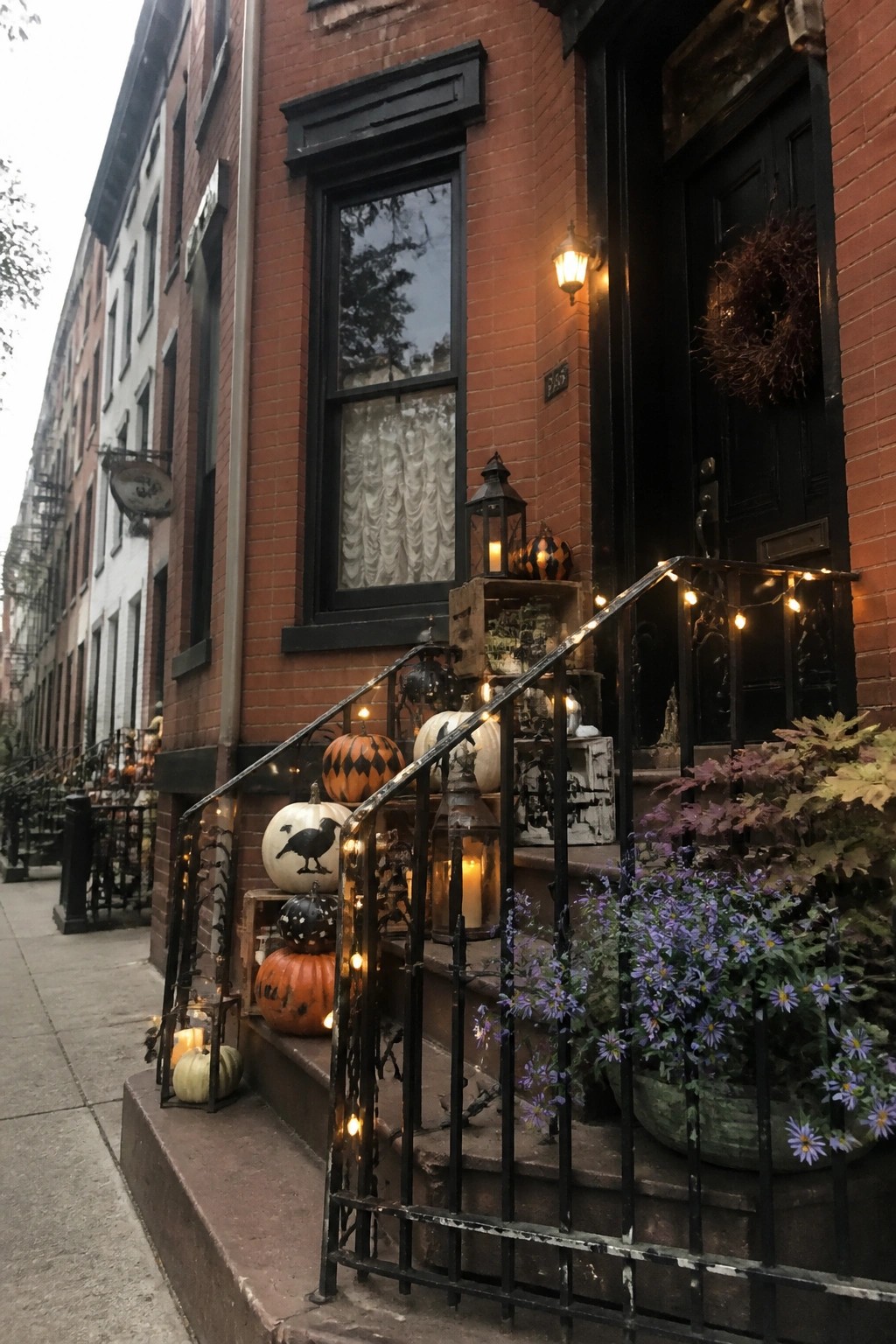 Halloween pumpkins, lanterns, and string lights adorn red brick brownstone stoop at dusk